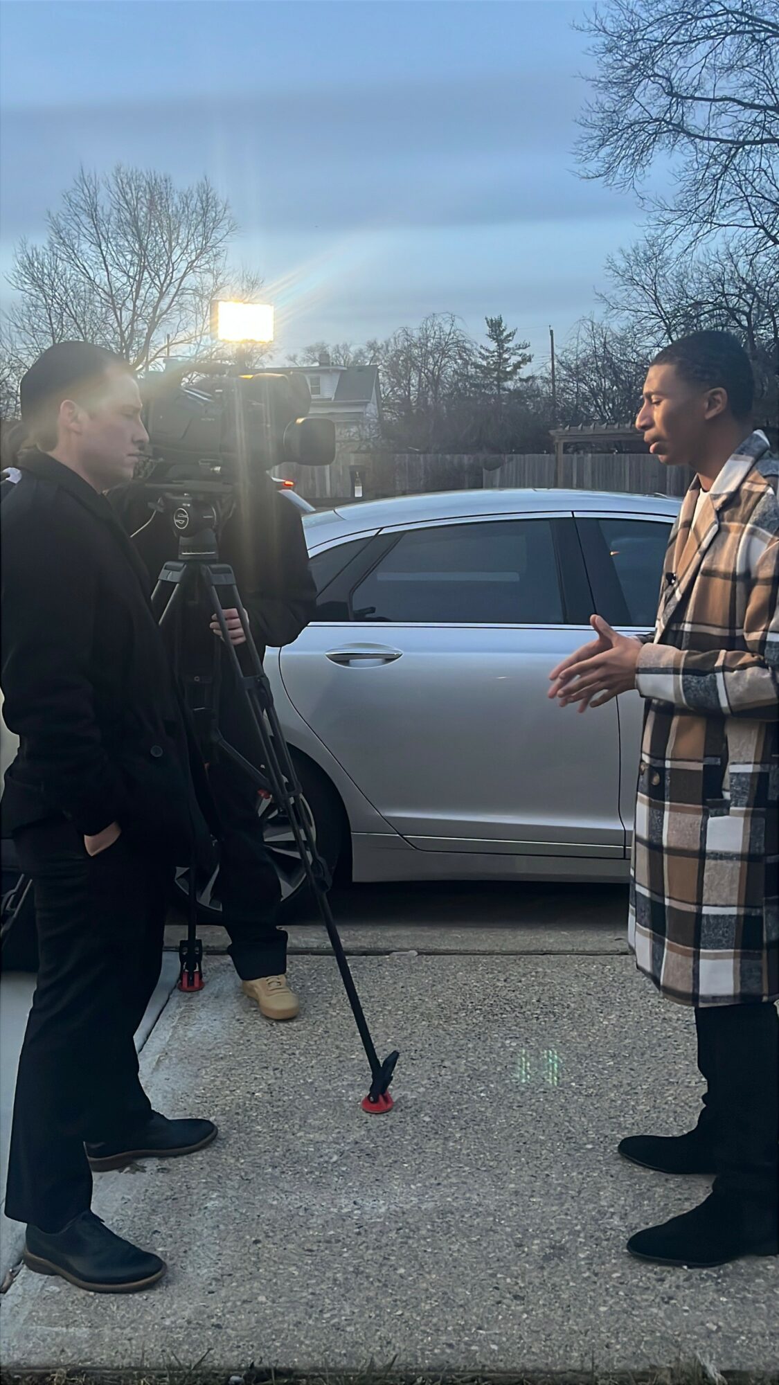 Two men are talking on a sidewalk near a parked car, with trees and a building in the background.