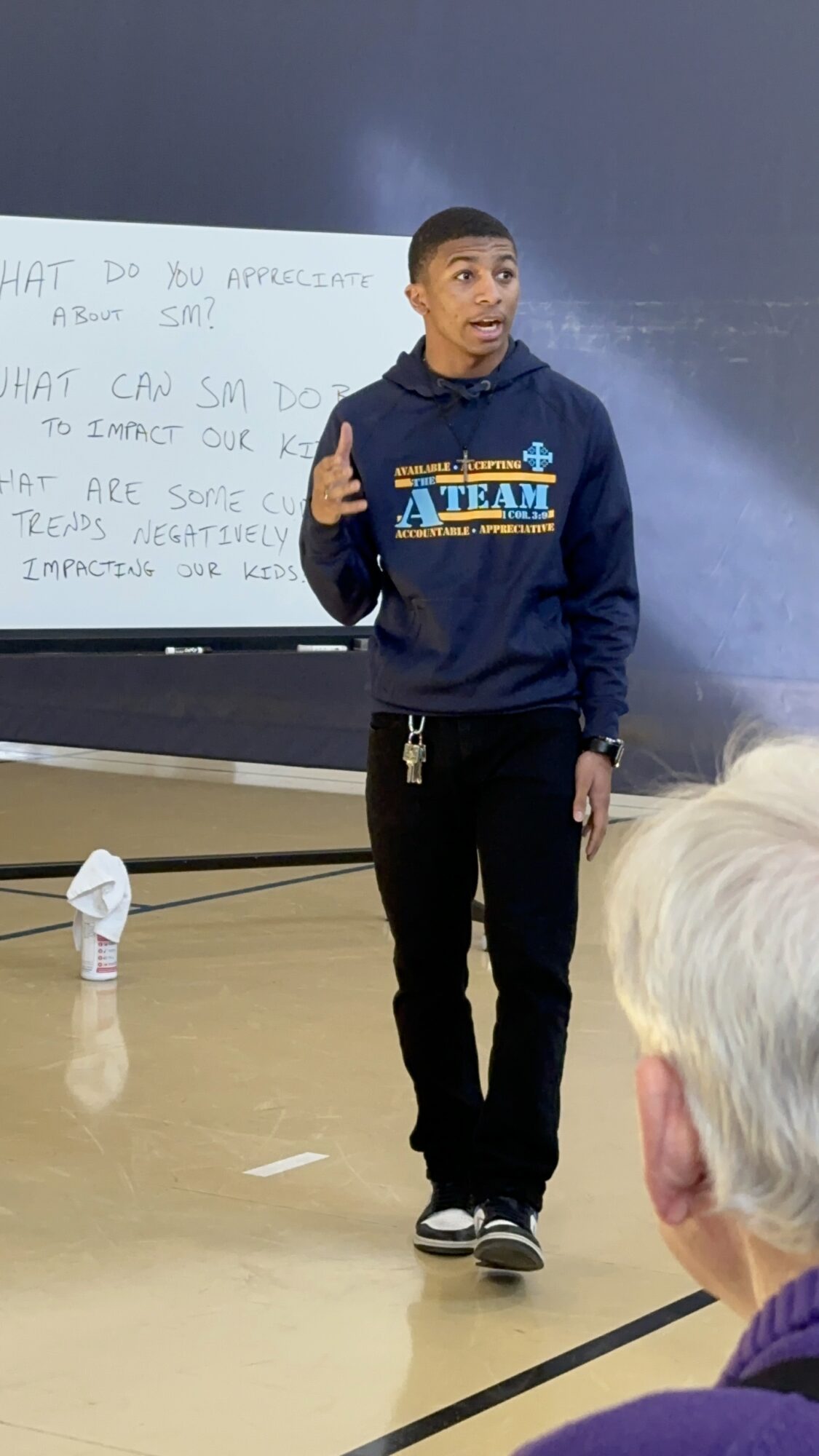 Young man standing and speaking in front of a classroom, with a whiteboard behind him and students listening.