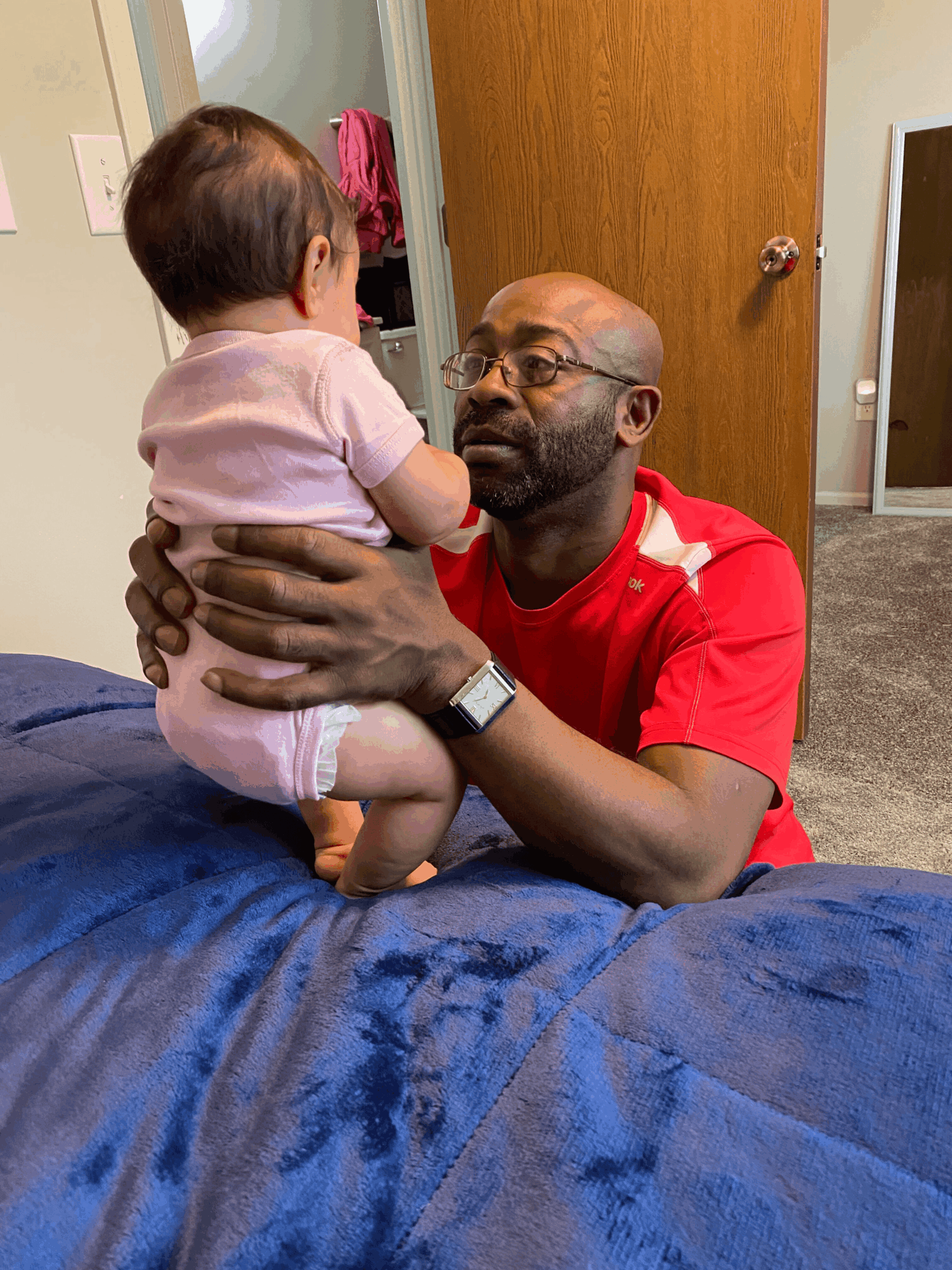 A man with glasses and a beard holds a young child in a pink shirt and shorts, indoors near a door.