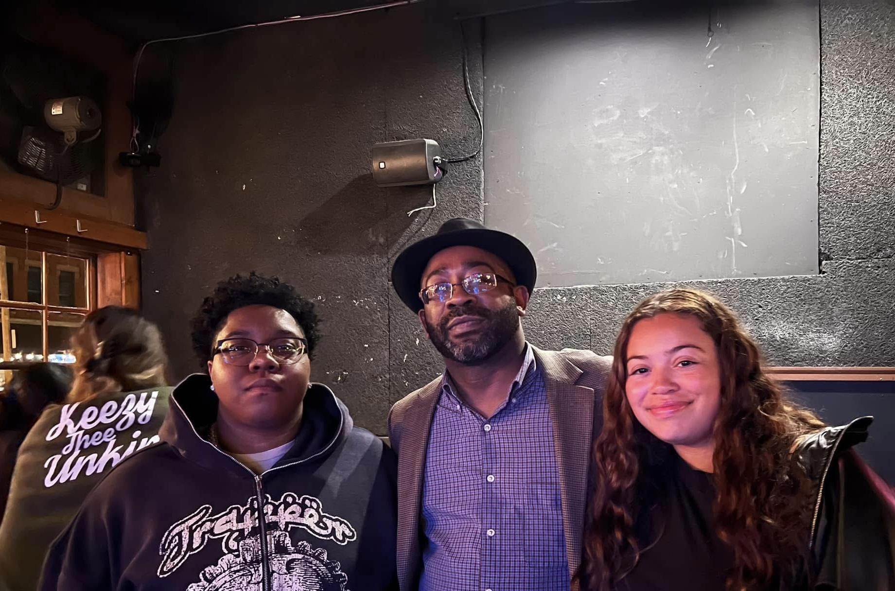 Three people standing together indoors, smiling, with a dark wall and a sign reading 'Keezy is the Unki' in the background.