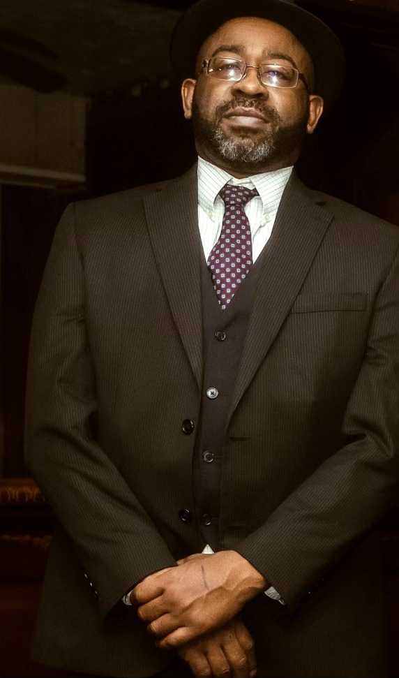 Man in formal suit with glasses and a hat, standing indoors with hands clasped.