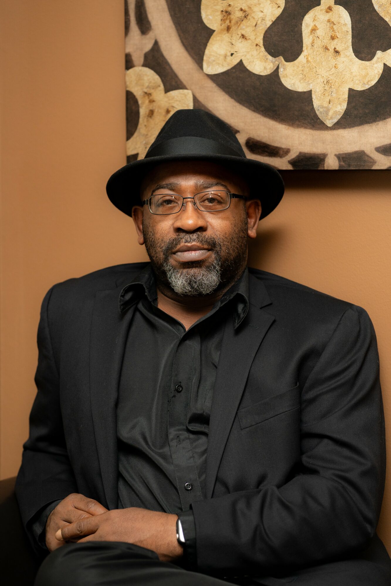 Man wearing a black hat and suit, standing in front of a decorative wall hanging, with a serious expression.