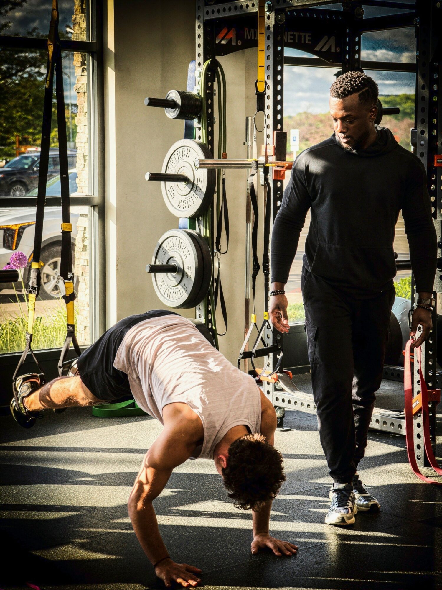 Person doing push-up on floor with a trainer observing in a gym, weightlifting equipment visible in background.