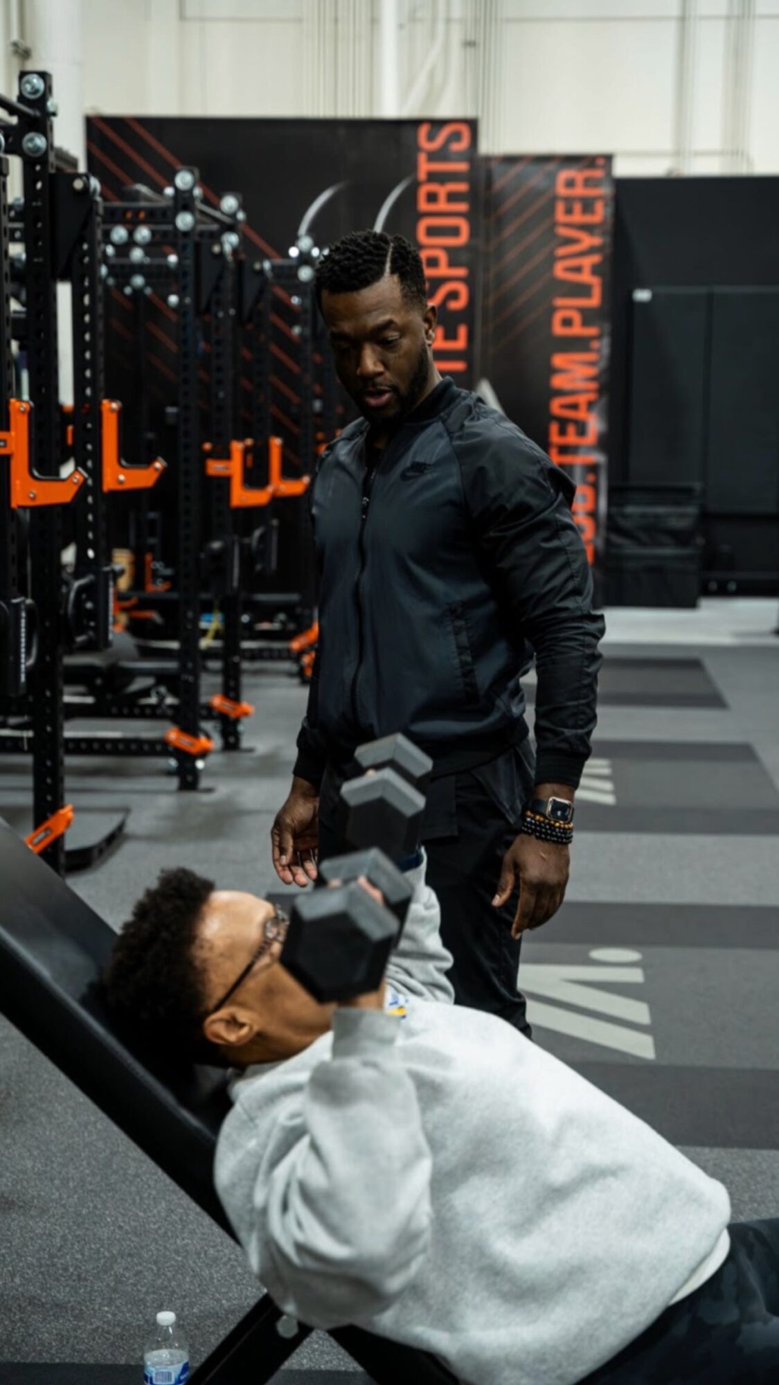 Two people in a gym, one sitting and drinking water, the other standing nearby, gym equipment in background.