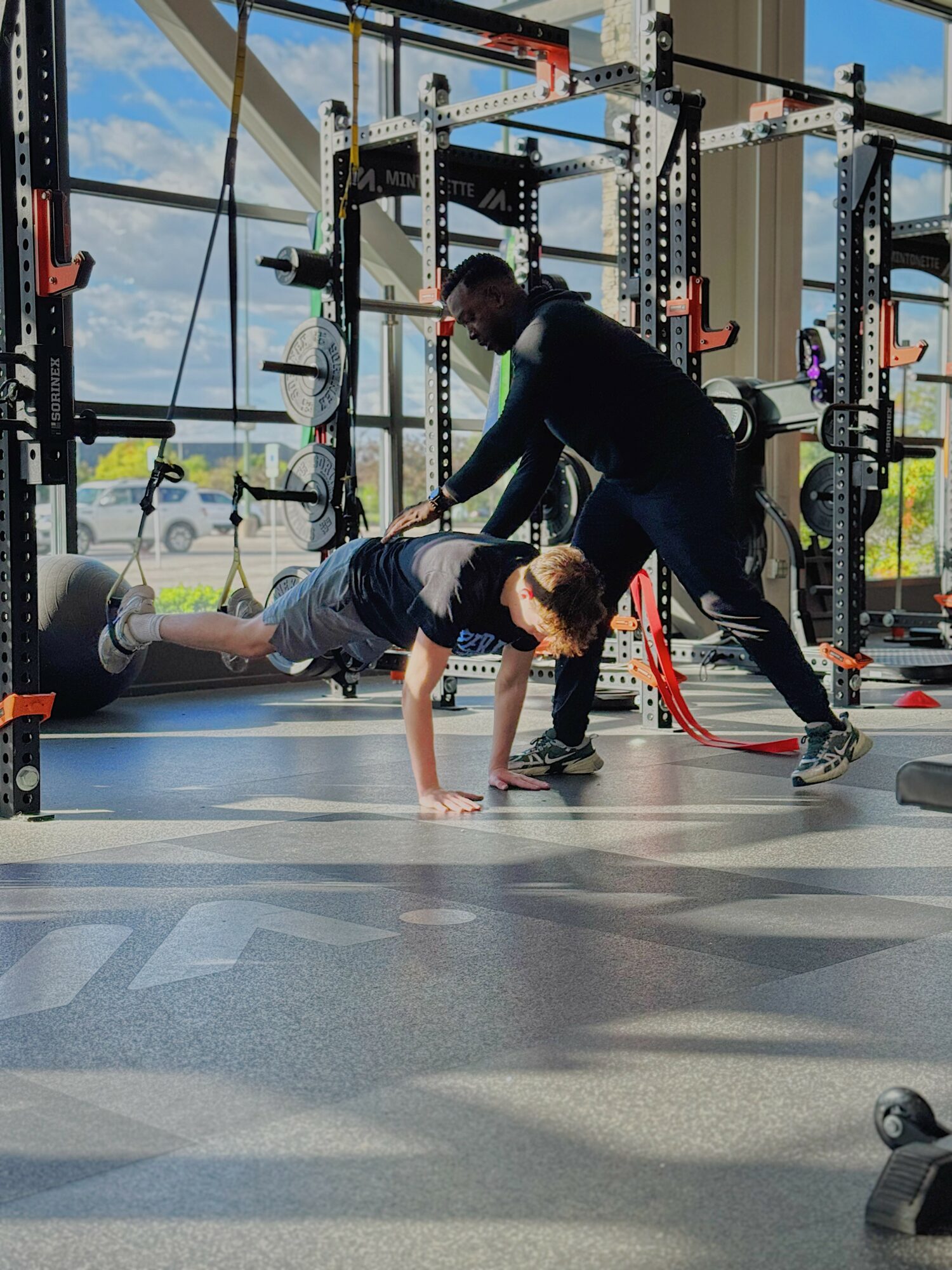 Two people exercising in a gym with large windows, one doing a plank and the other assisting, gym equipment visible.