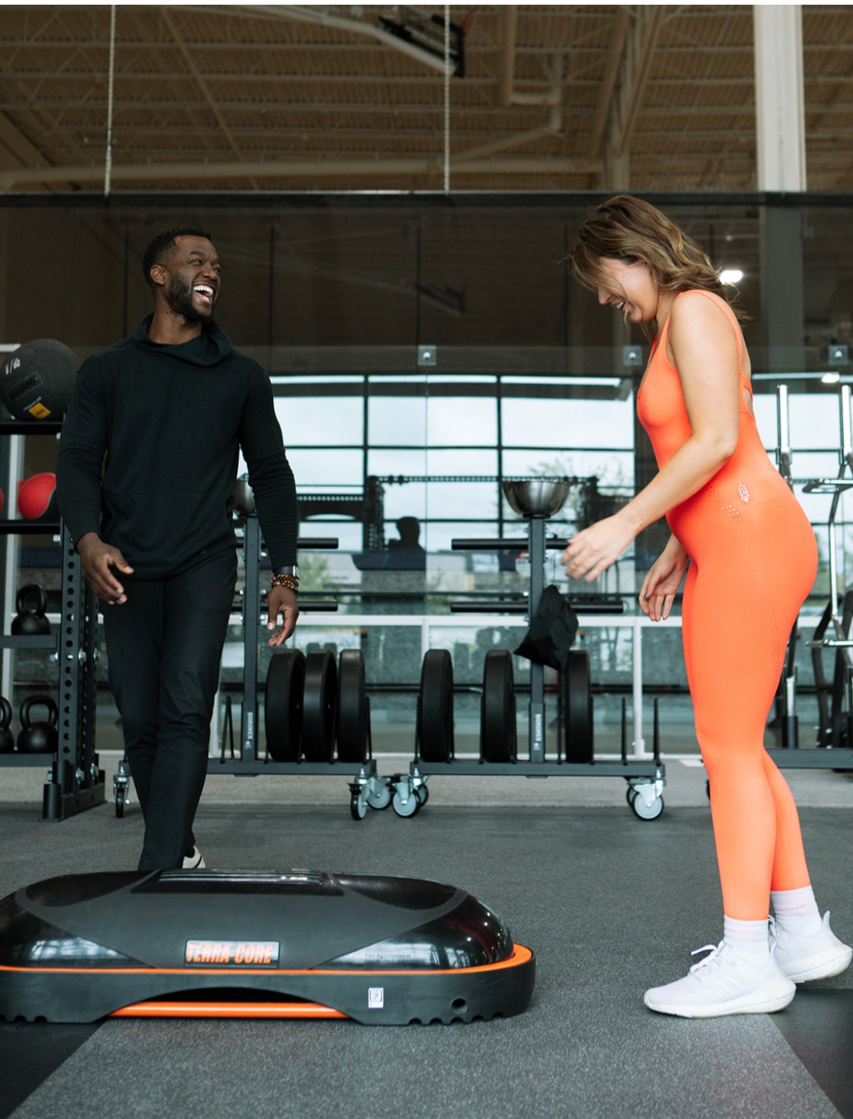 Two people in a gym, one woman in orange workout clothes and white shoes, the other man in black, smiling.