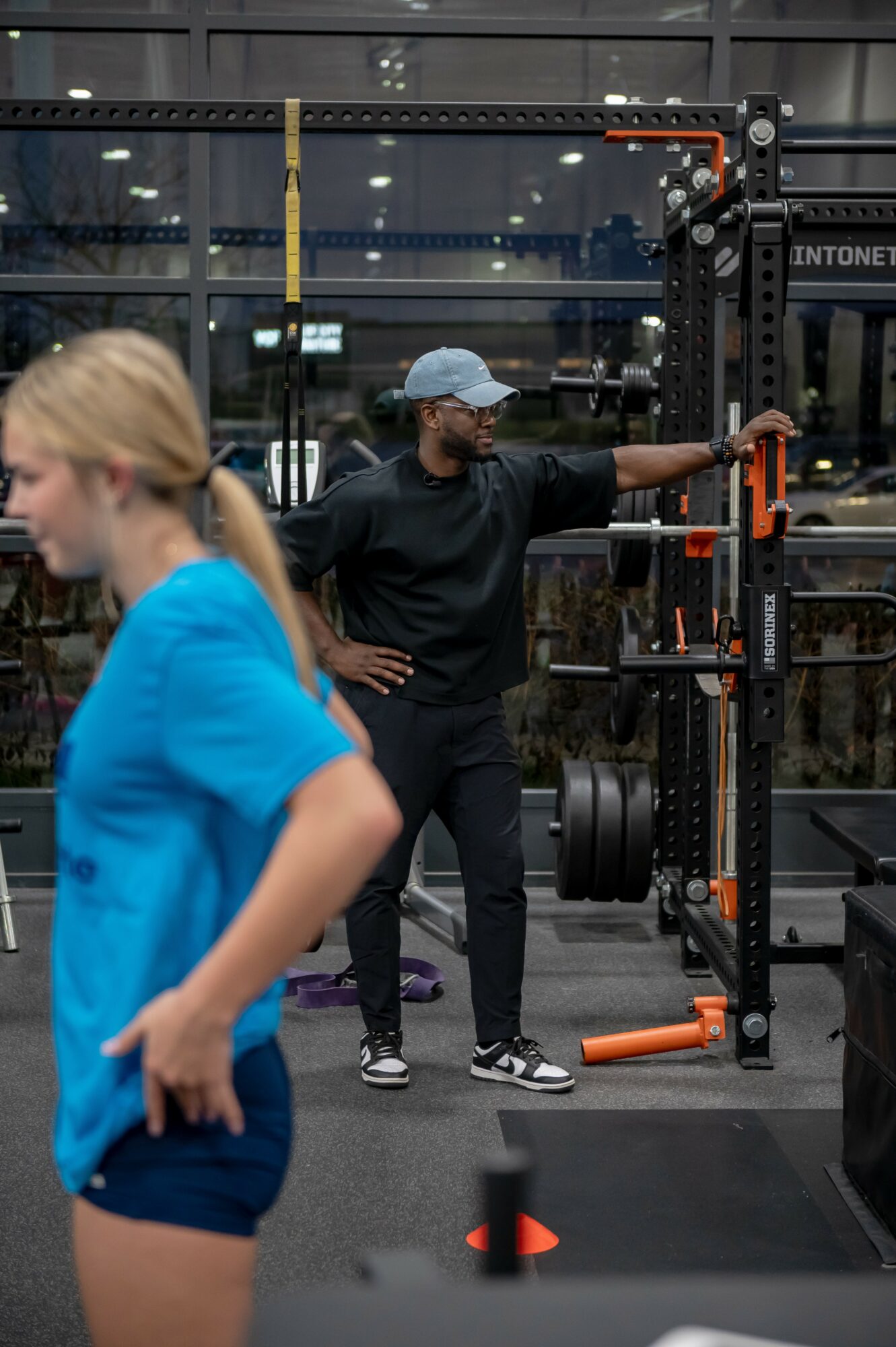 Person in black shirt and cap standing next to gym equipment, with a woman in blue in foreground, inside a gym.