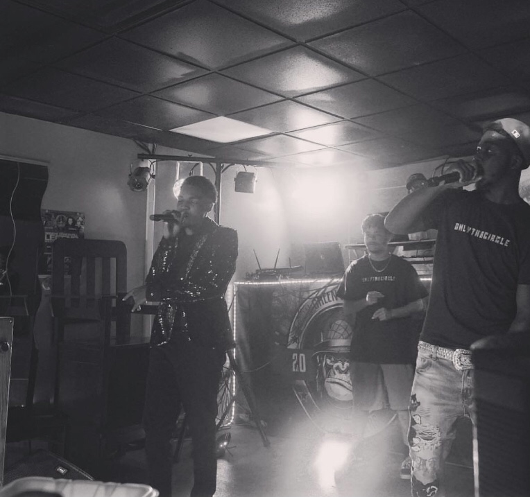 Three people in a dimly lit room, one with a microphone, another with a cap, and a third with glasses, standing near a DJ setup.