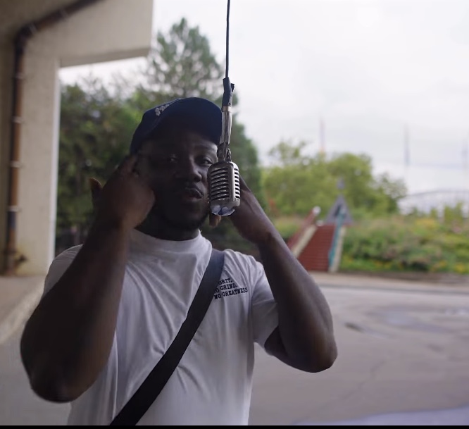 Person wearing a white shirt and cap, holding a radio antenna outdoors, with trees and a road in the background.