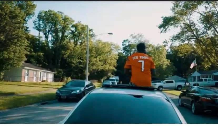Person sitting on a car with a sports jersey, in a parking lot with trees and houses in the background.