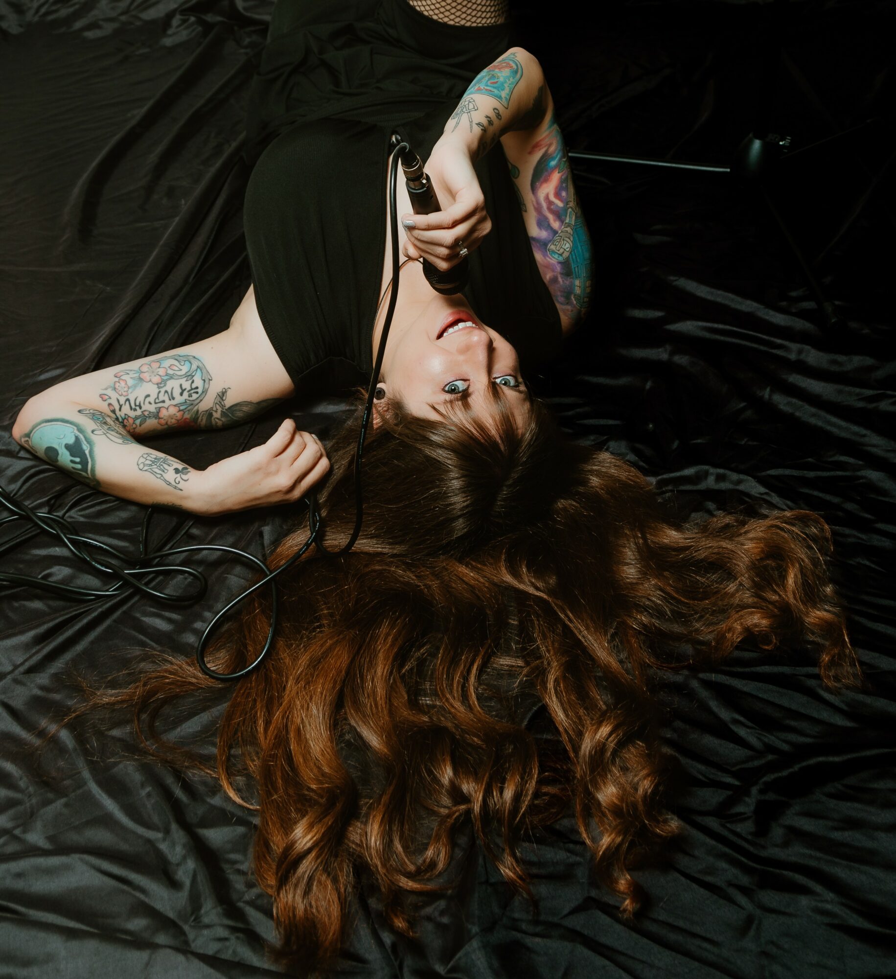 Woman with long hair and tattoos lying on black surface, holding a microphone, head upside down, hair spread out.