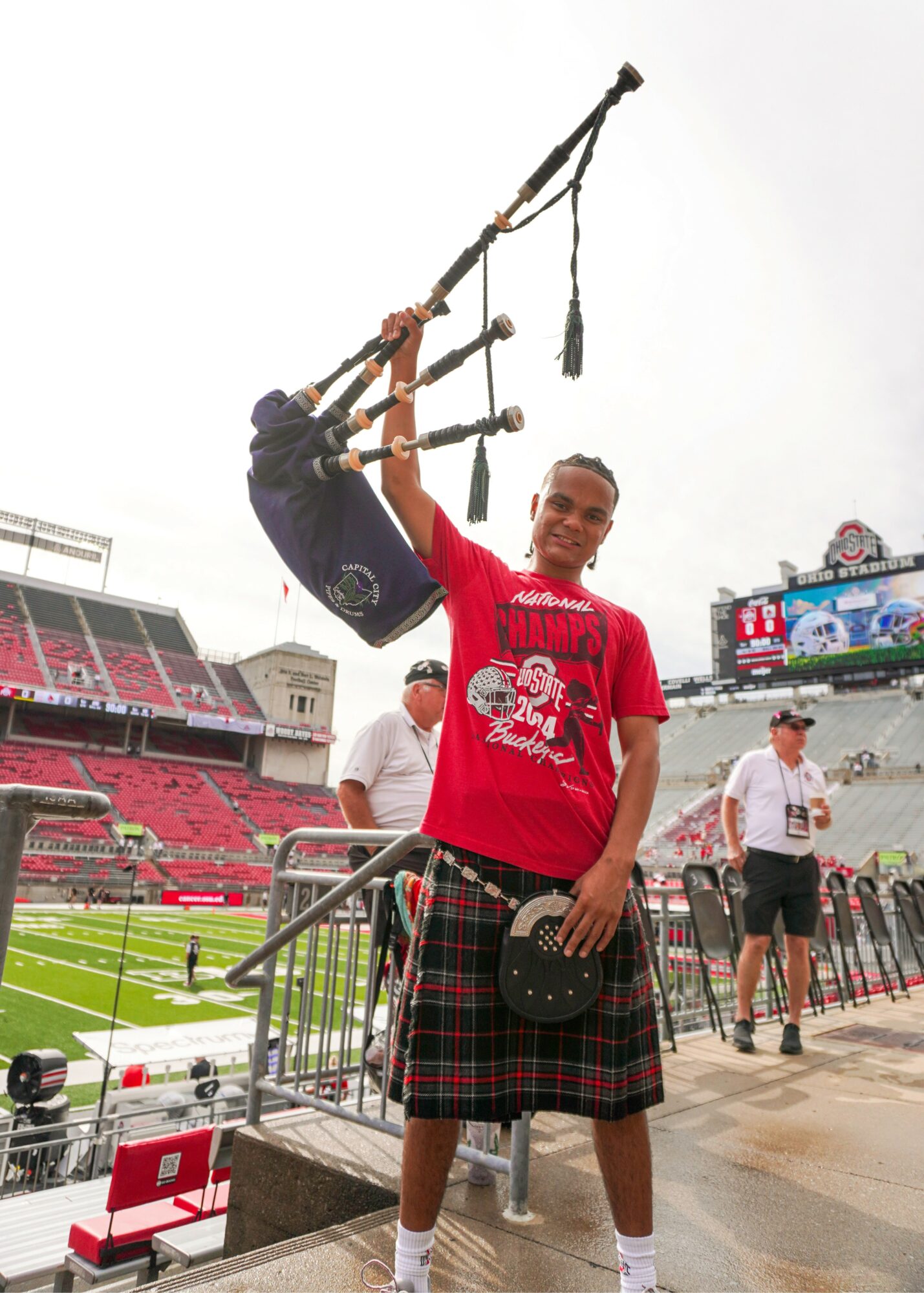 Person in red shirt and plaid kilt holding a bag, standing in a stadium with people and seating in background.
