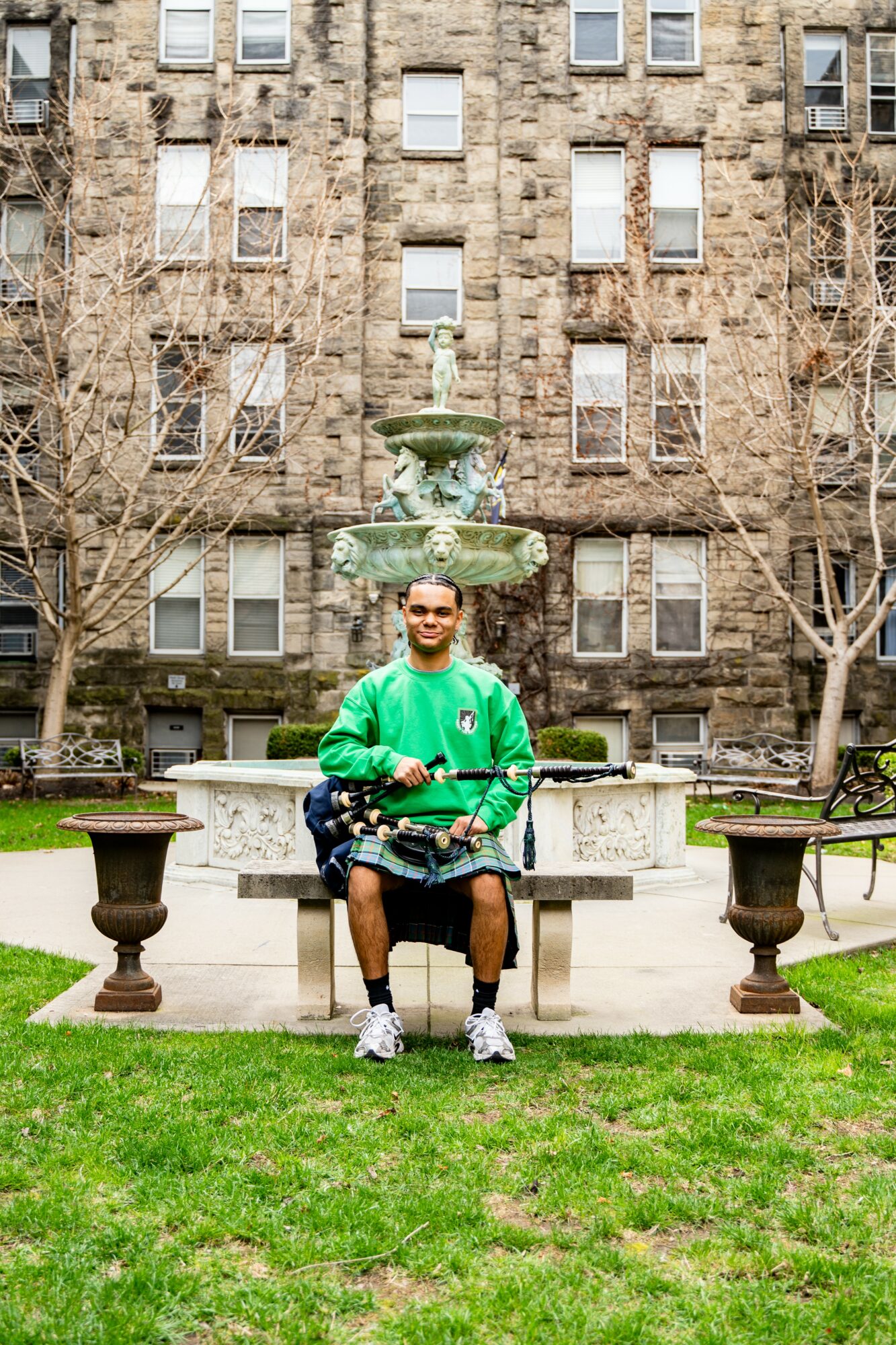 Person sitting on a bench in front of a fountain with a stone building and leafless trees behind.
