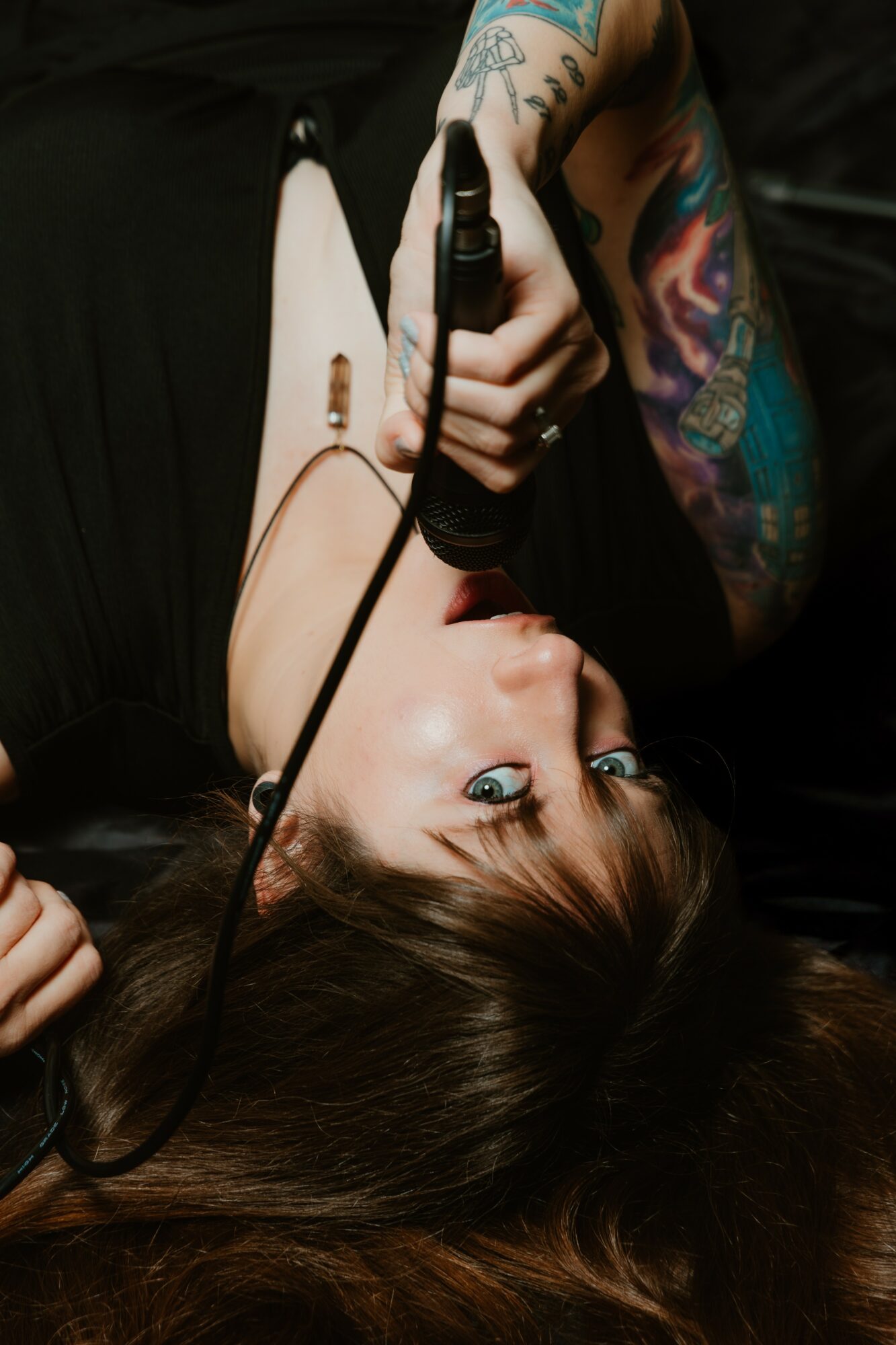 Woman with tattoos lying down, holding a microphone, with long brown hair and colorful tattoos on her arm.