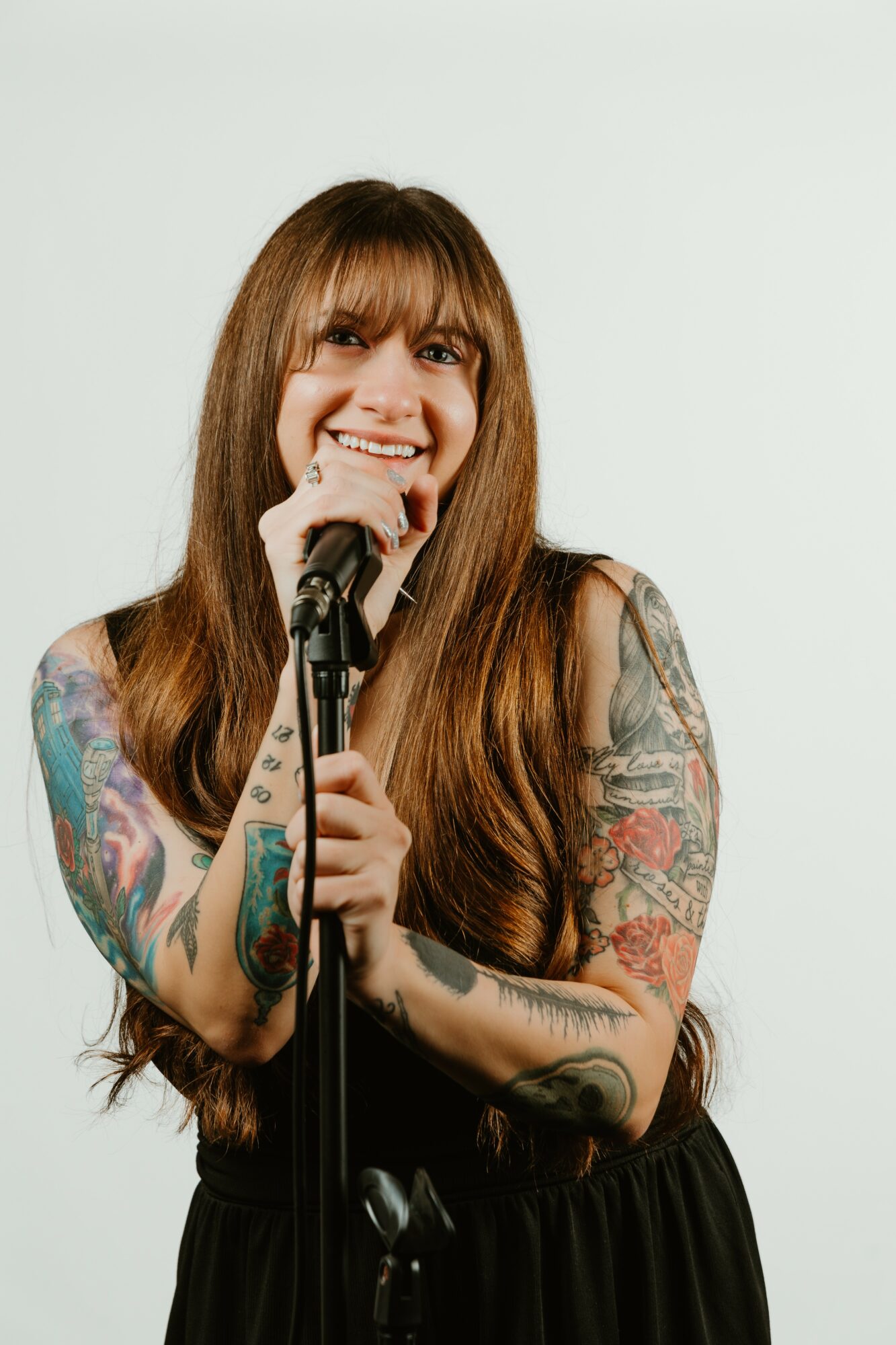 Woman with long hair and tattoos smiling, holding a microphone, against a plain background.