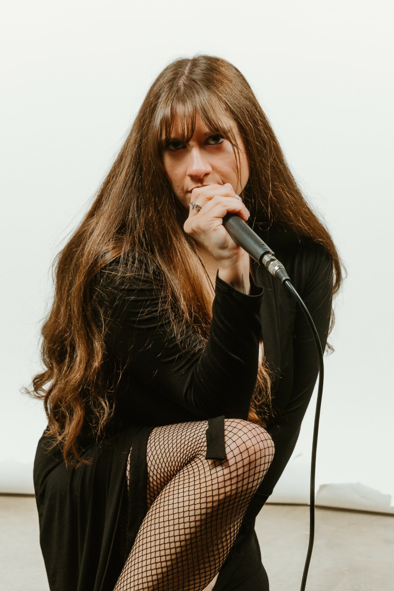 Woman with long hair singing into a microphone, wearing a black outfit and fishnet stockings, against a plain background.