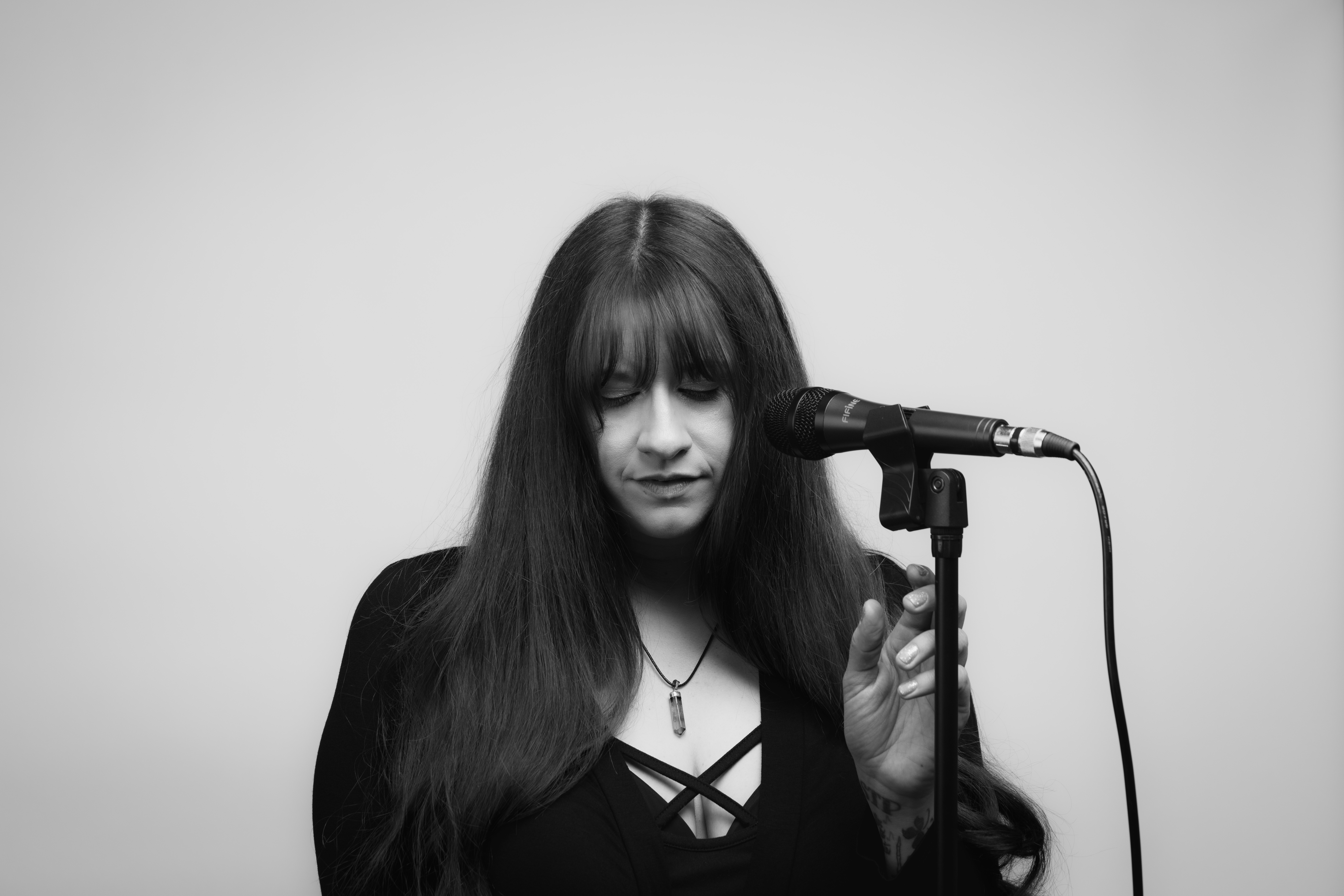 Woman with long dark hair and bangs speaking into a microphone against a plain background.