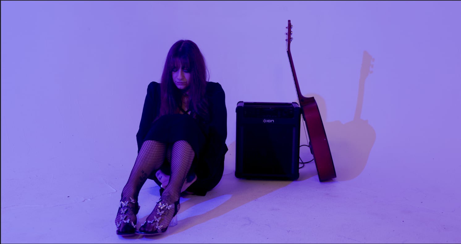 Woman sitting on the floor with two guitars and an amplifier, purple background, facing forward.
