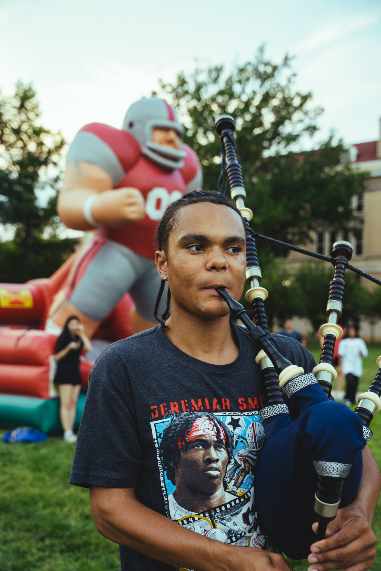 Young man playing bagpipes outdoors with a large football player statue in the background, trees, and people in a park.