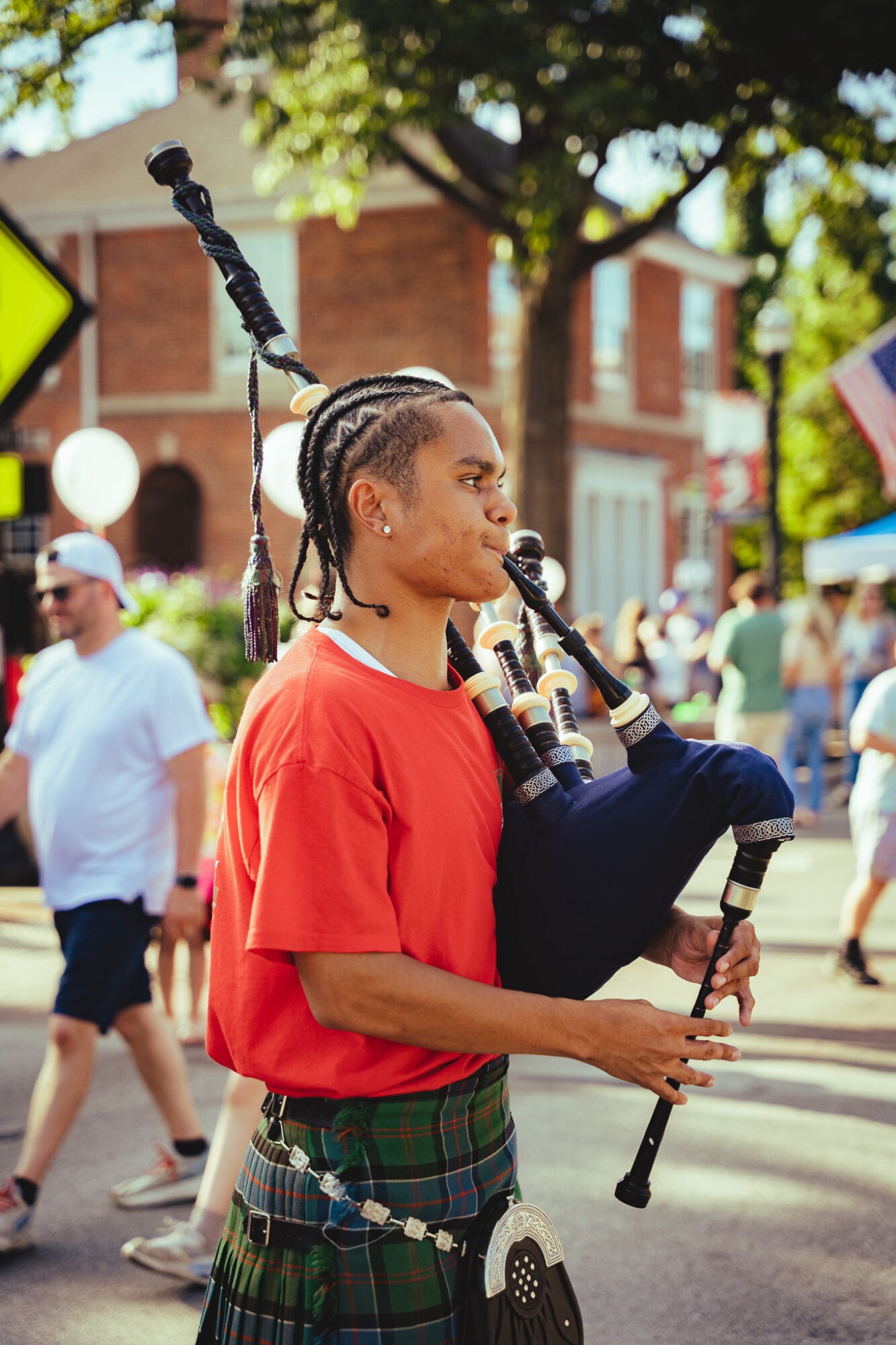 Woman playing bagpipes in a street festival with people and buildings in the background.