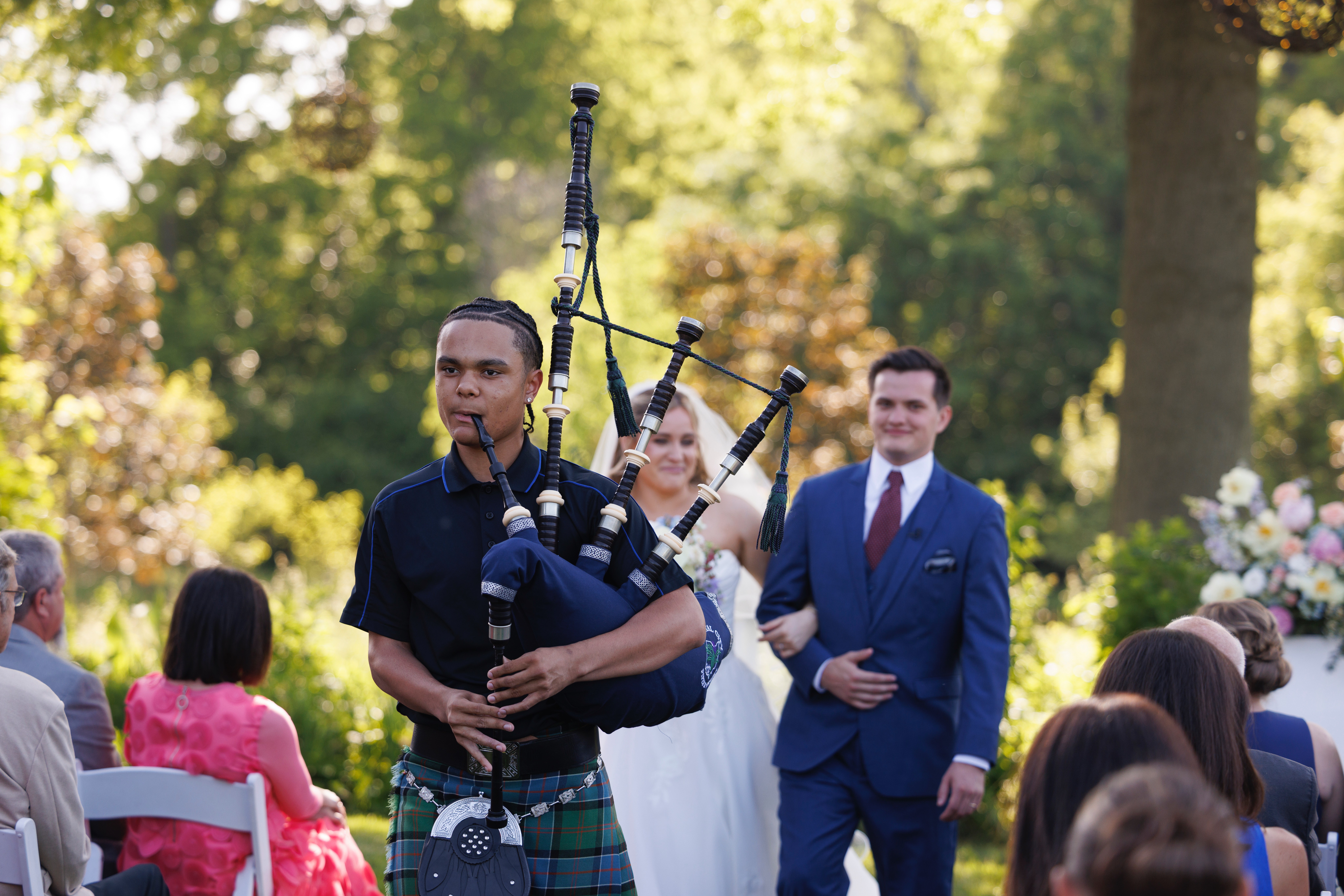 Person playing bagpipes outdoors with a group of people watching, trees and sunlight in background.