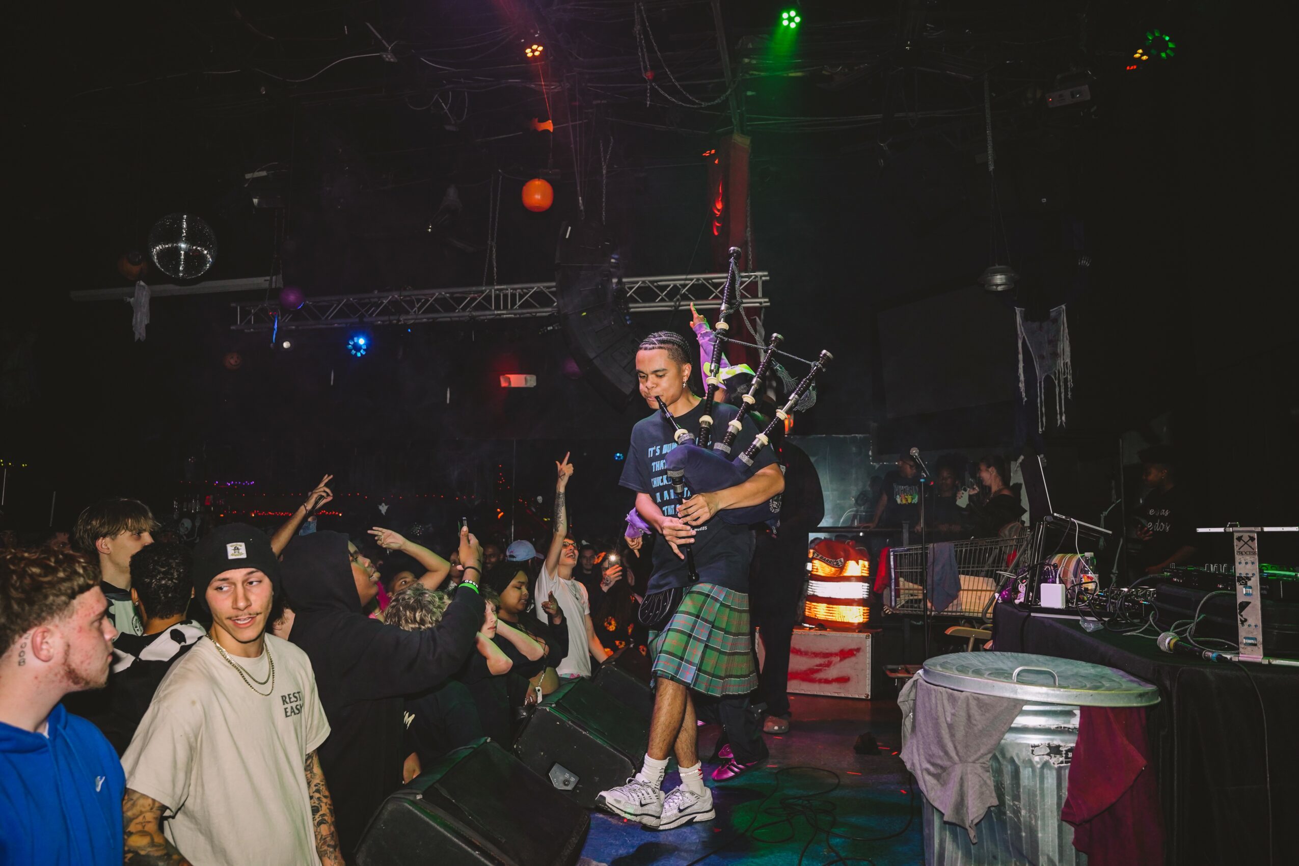 Person playing bagpipes on stage with audience in background, stage lights visible, dark setting, lively atmosphere.