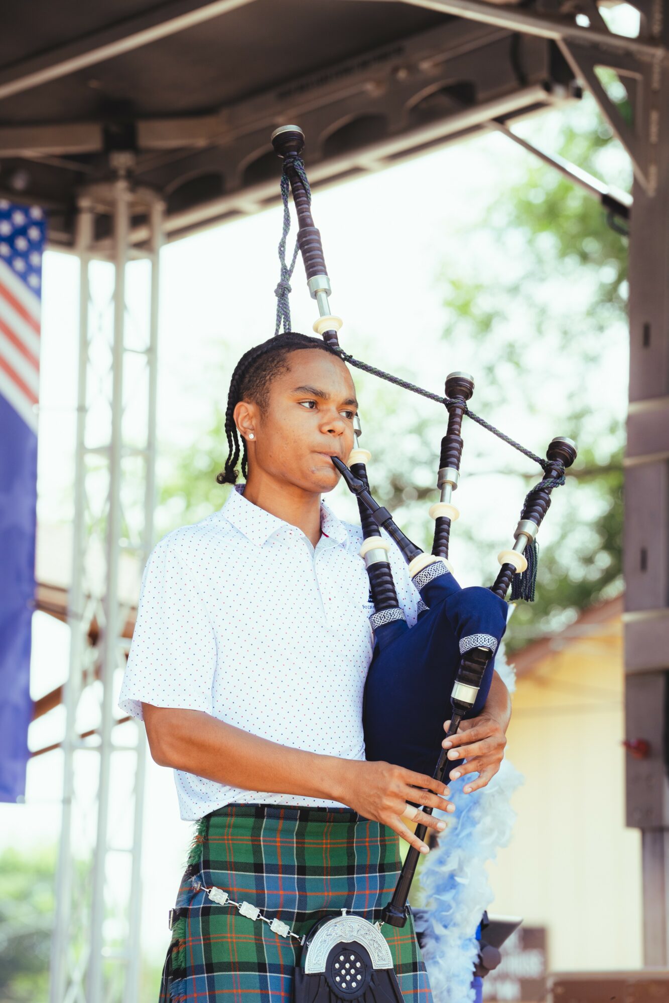 Young man playing bagpipes outdoors with a blurred background, American flag, and metal structure visible.