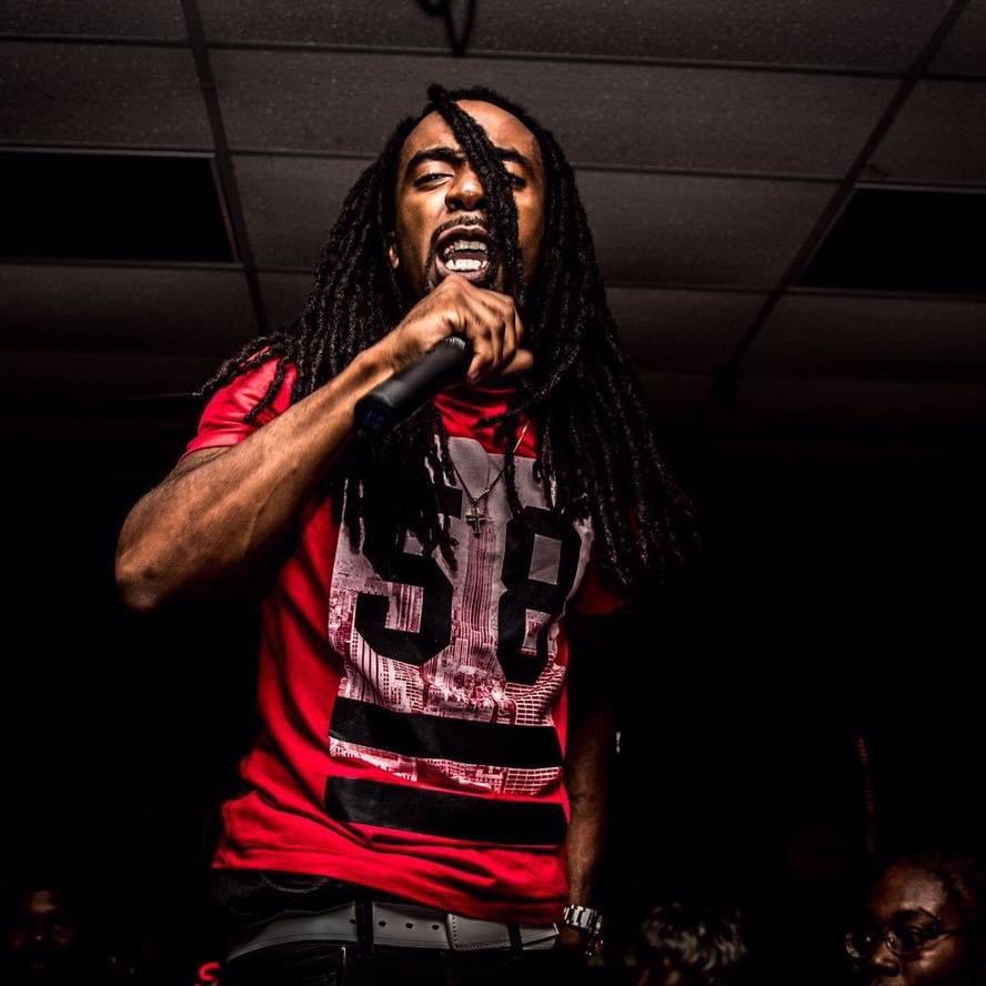 Person with long dreadlocks singing into a microphone, wearing a red and black shirt, on stage with a dark background.