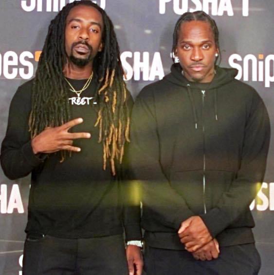Two men standing side by side in front of a backdrop with text, one making a peace sign, both looking at the camera.