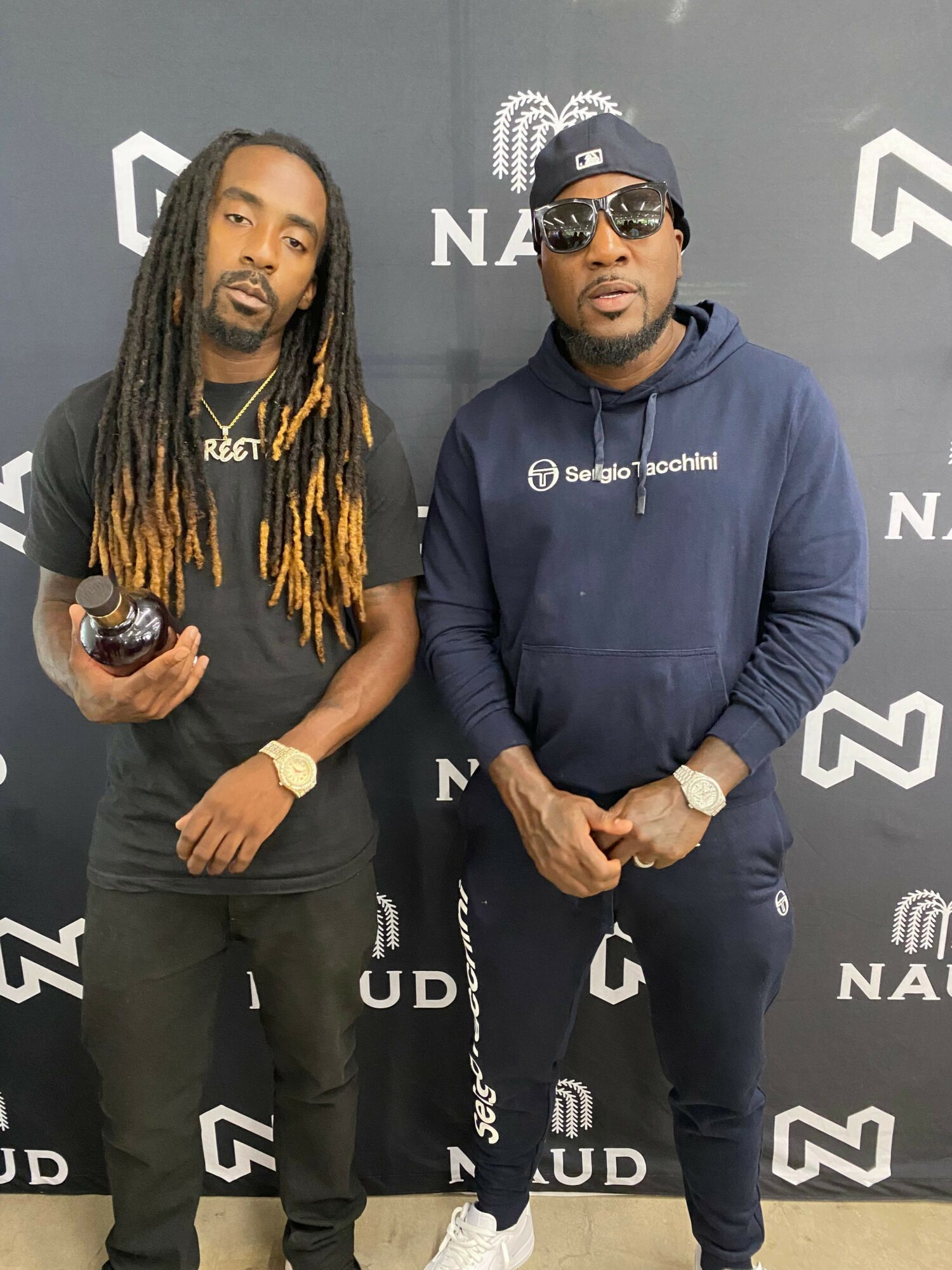 Two people standing side by side, one wearing a dark hoodie and the other with long dreadlocks, against a gray background with logos.