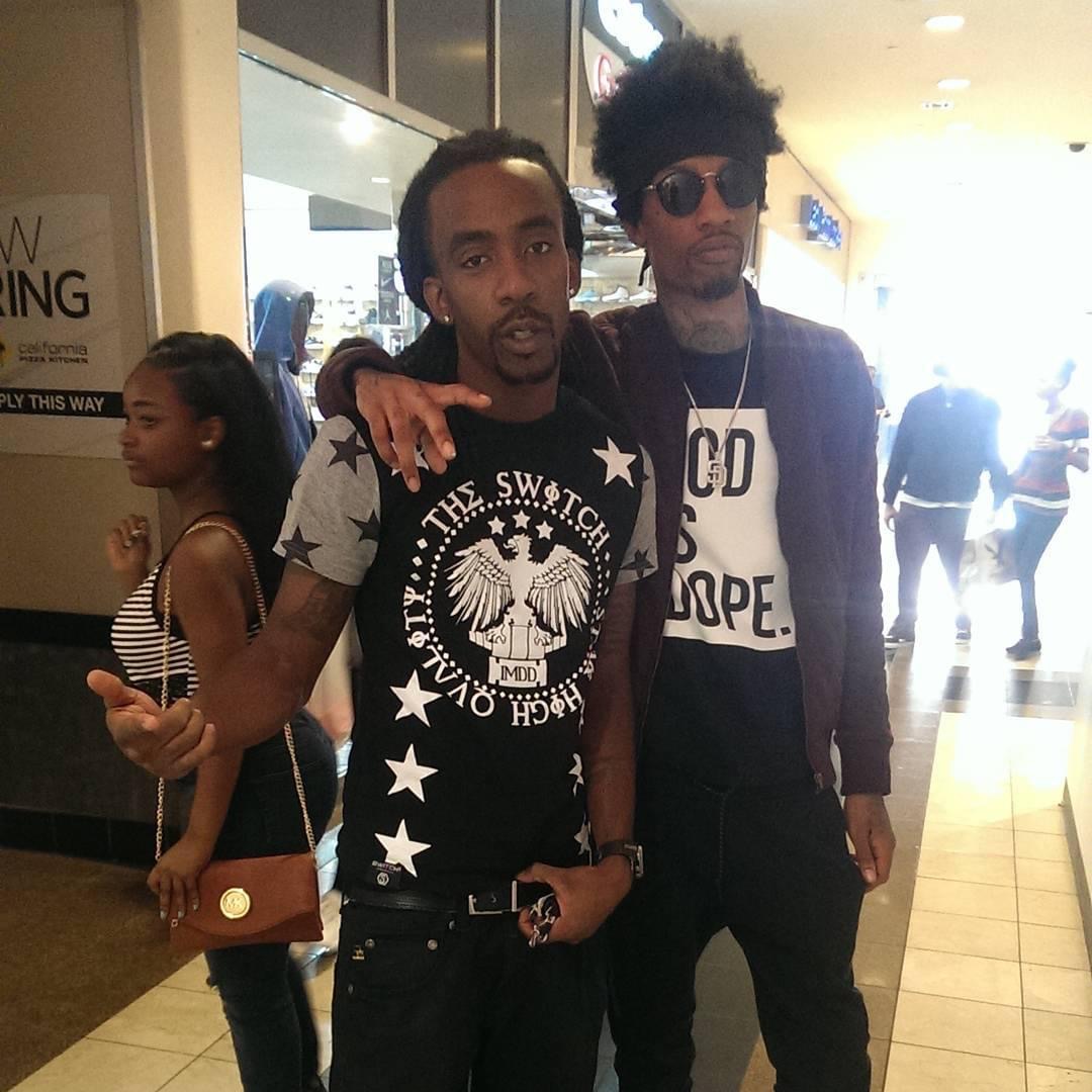 Two men standing together in a shopping mall, one with sunglasses and the other with a graphic t-shirt, people in background.
