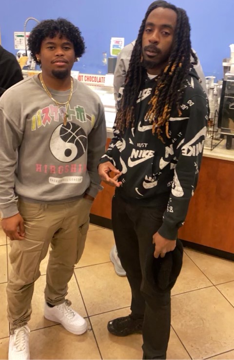 Two young men standing indoors, one with curly hair wearing a gray sweatshirt, the other with long dreadlocks wearing a black Nike hoodie.