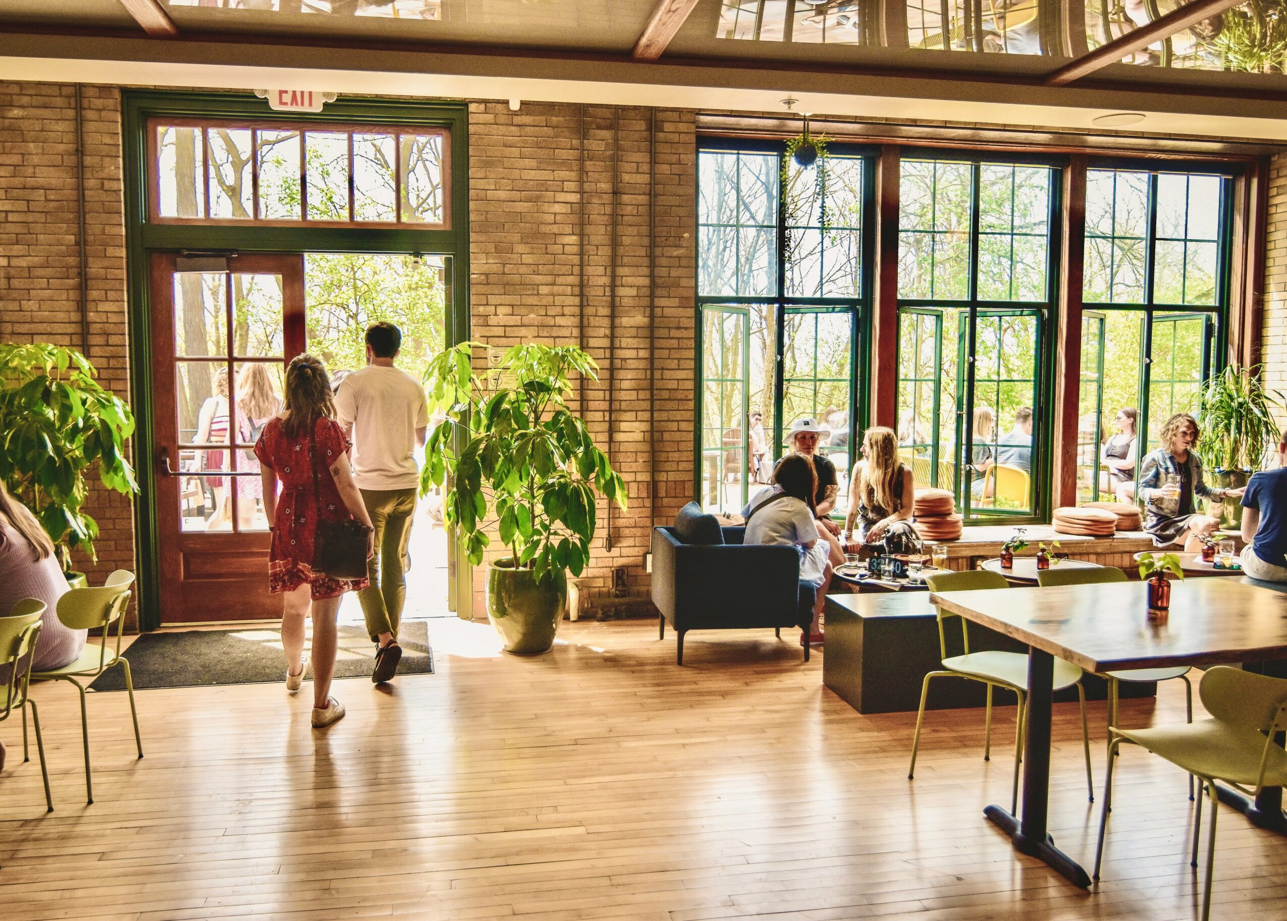 People walking into a bright, spacious cafe with large windows, plants, and seating areas, during daytime.
