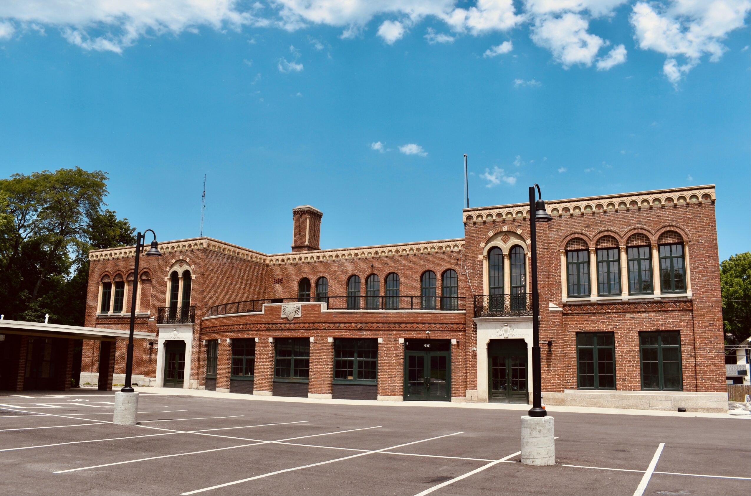 Brick building with arched windows and a parking lot in front, under a blue sky with clouds.