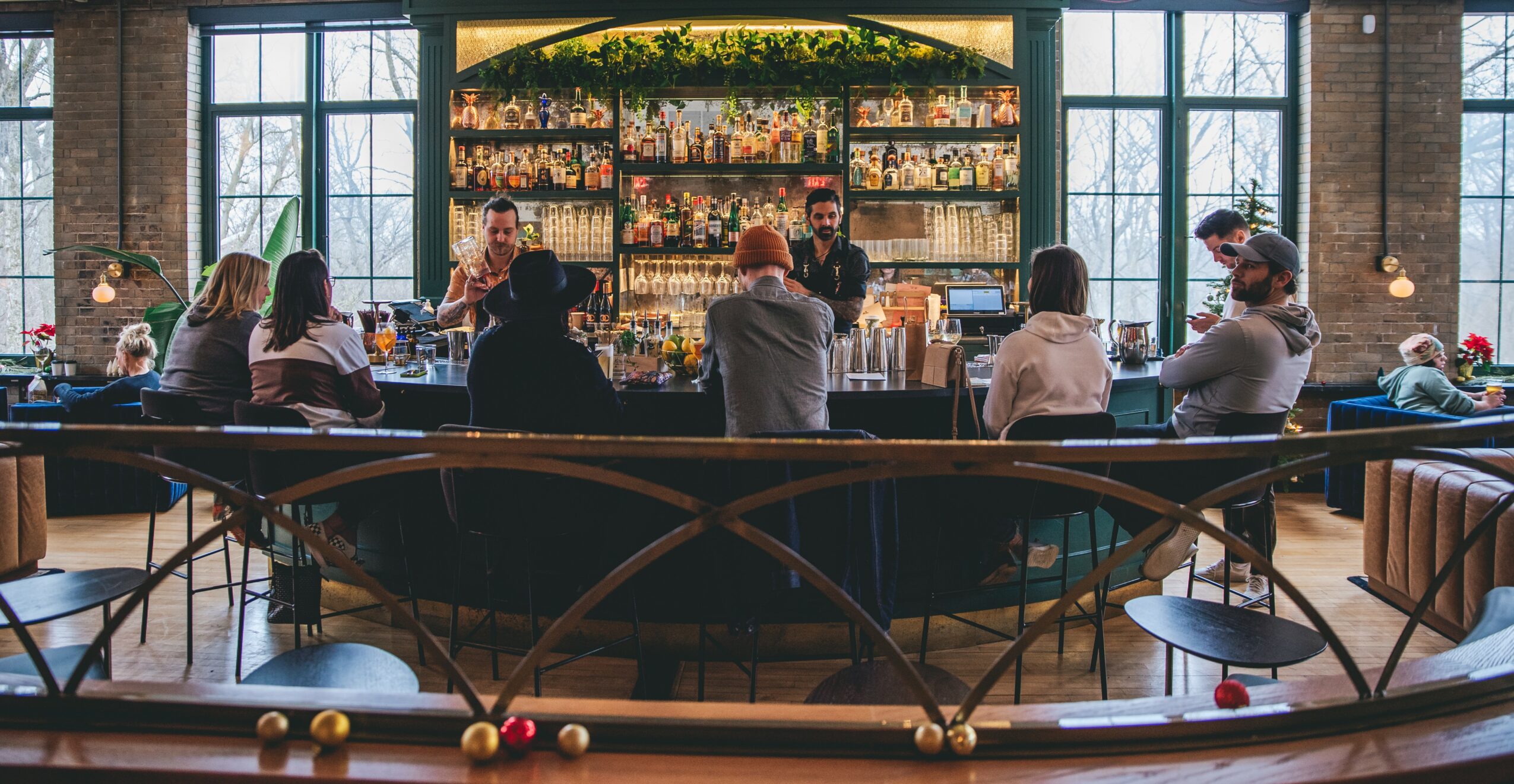 People sitting at a bar with large windows and shelves of bottles behind the bartender, in a spacious, well-lit setting.