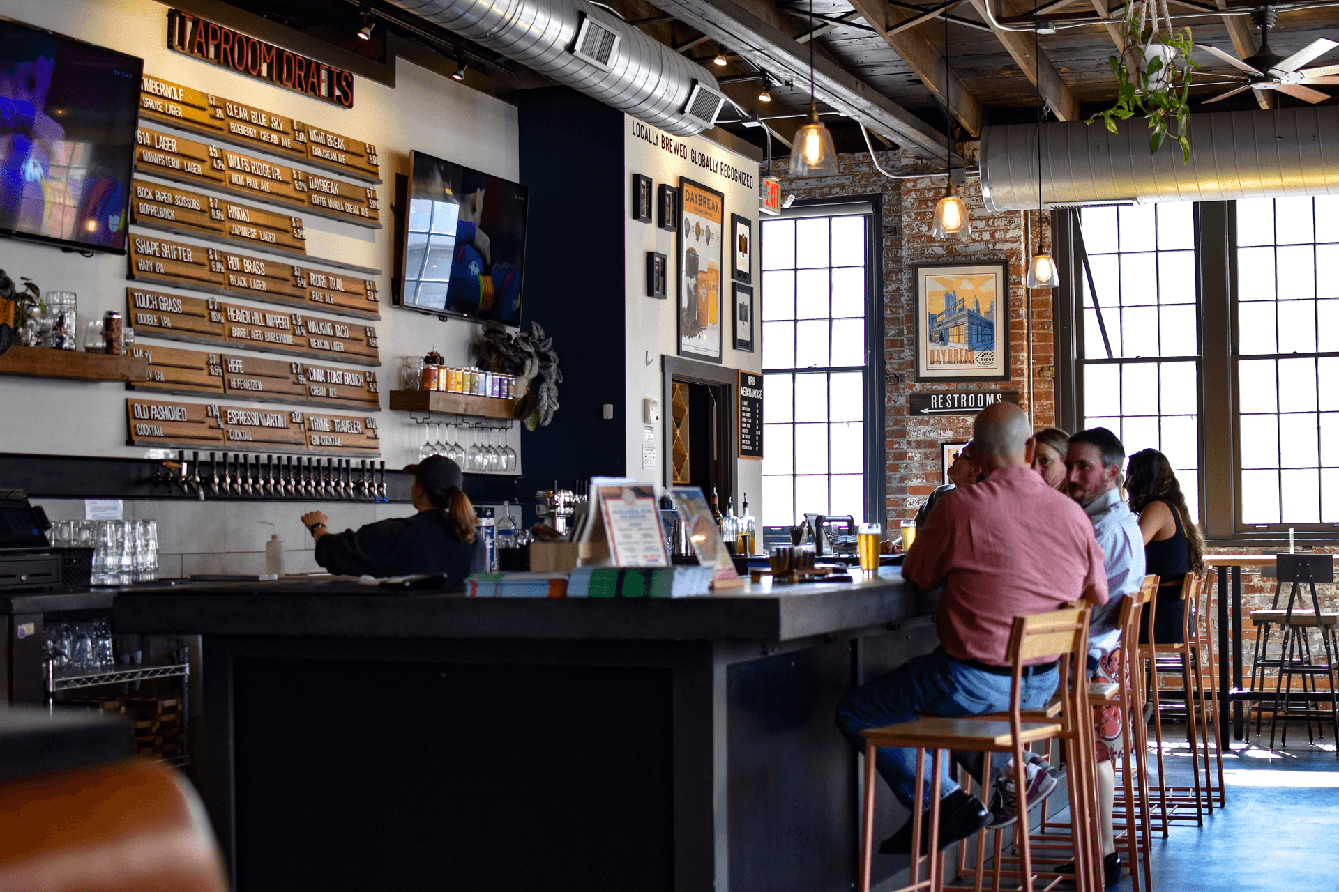 Interior of a modern cafe with large windows, bar counter, and people sitting and chatting, with shelves and artwork on walls.