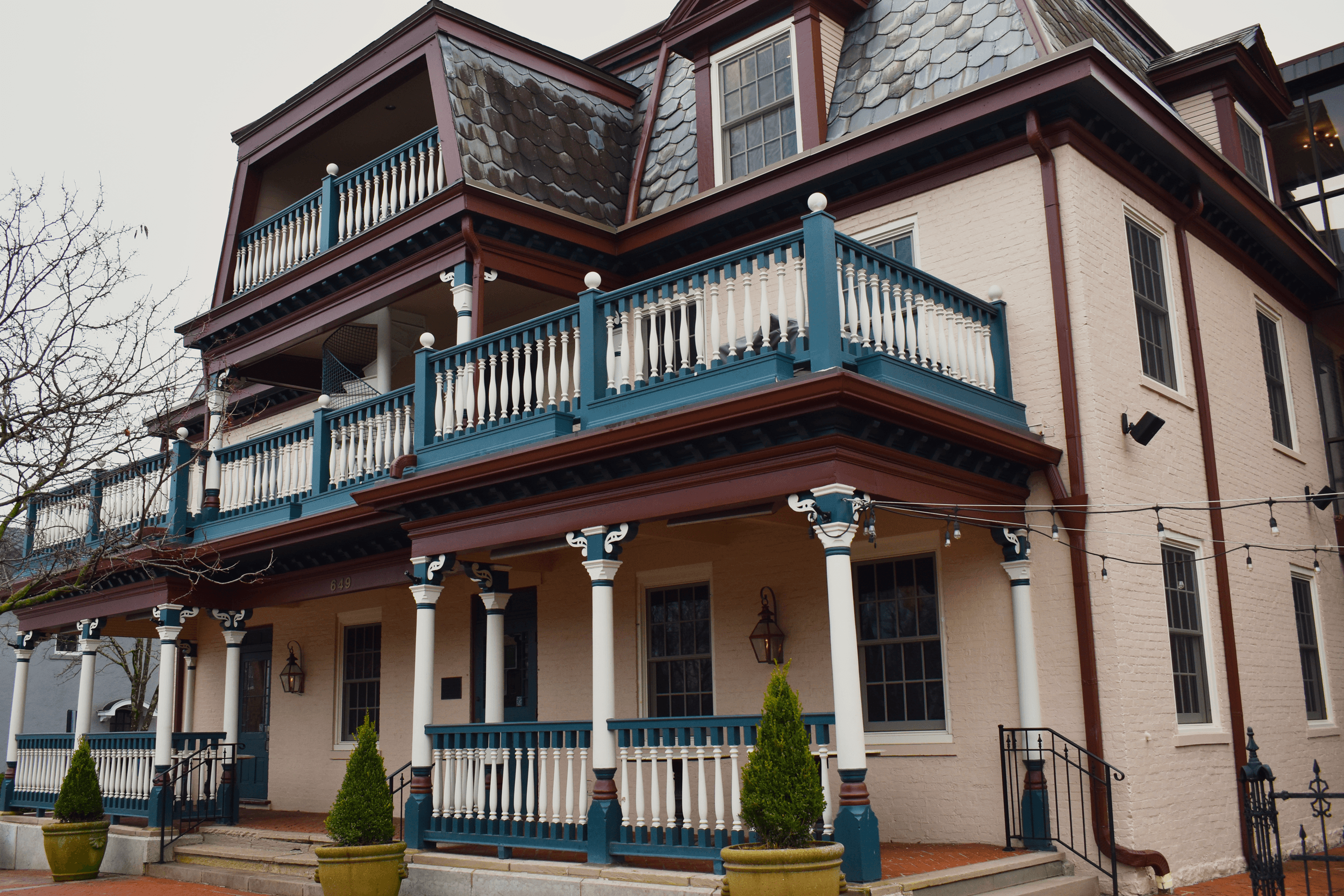 Multi-story house with balconies, columns, and decorative railings, featuring a beige exterior with blue accents and a sloped roof.