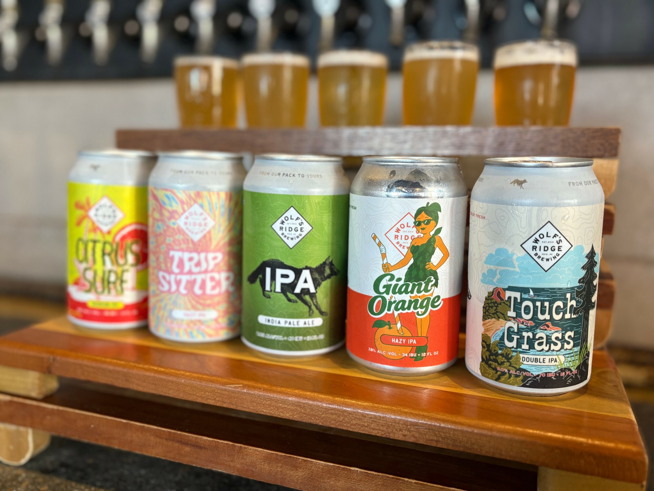 Six cans of craft beer on a wooden tray with glasses of beer in the background.