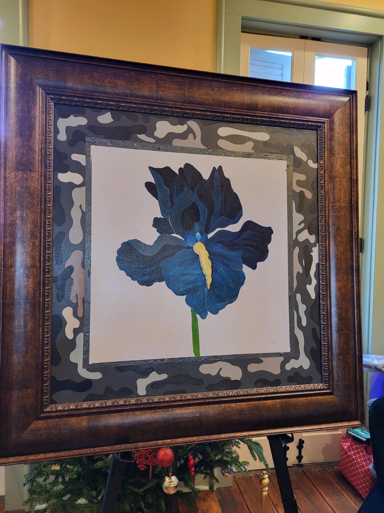 Framed painting of a blue flower with yellow center on a white background, with decorative border, leaning against a wall.