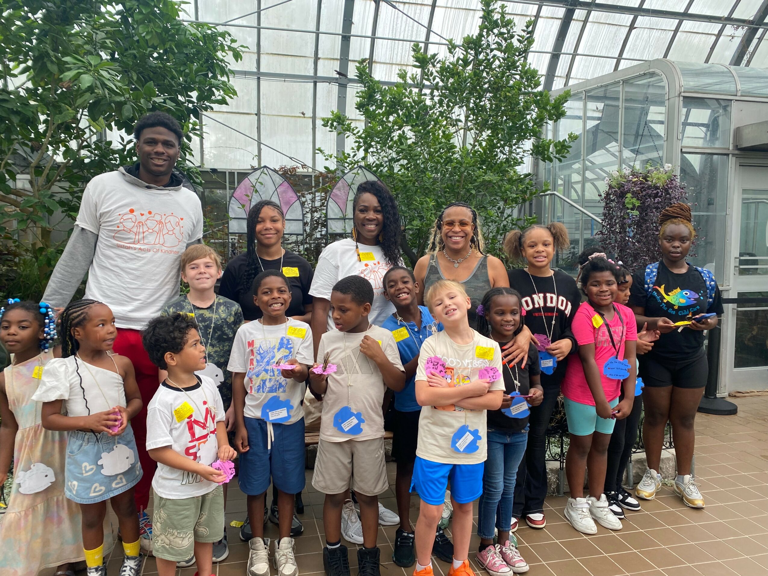 Group of children and adults standing inside a greenhouse with plants and glass walls, some holding colorful items.