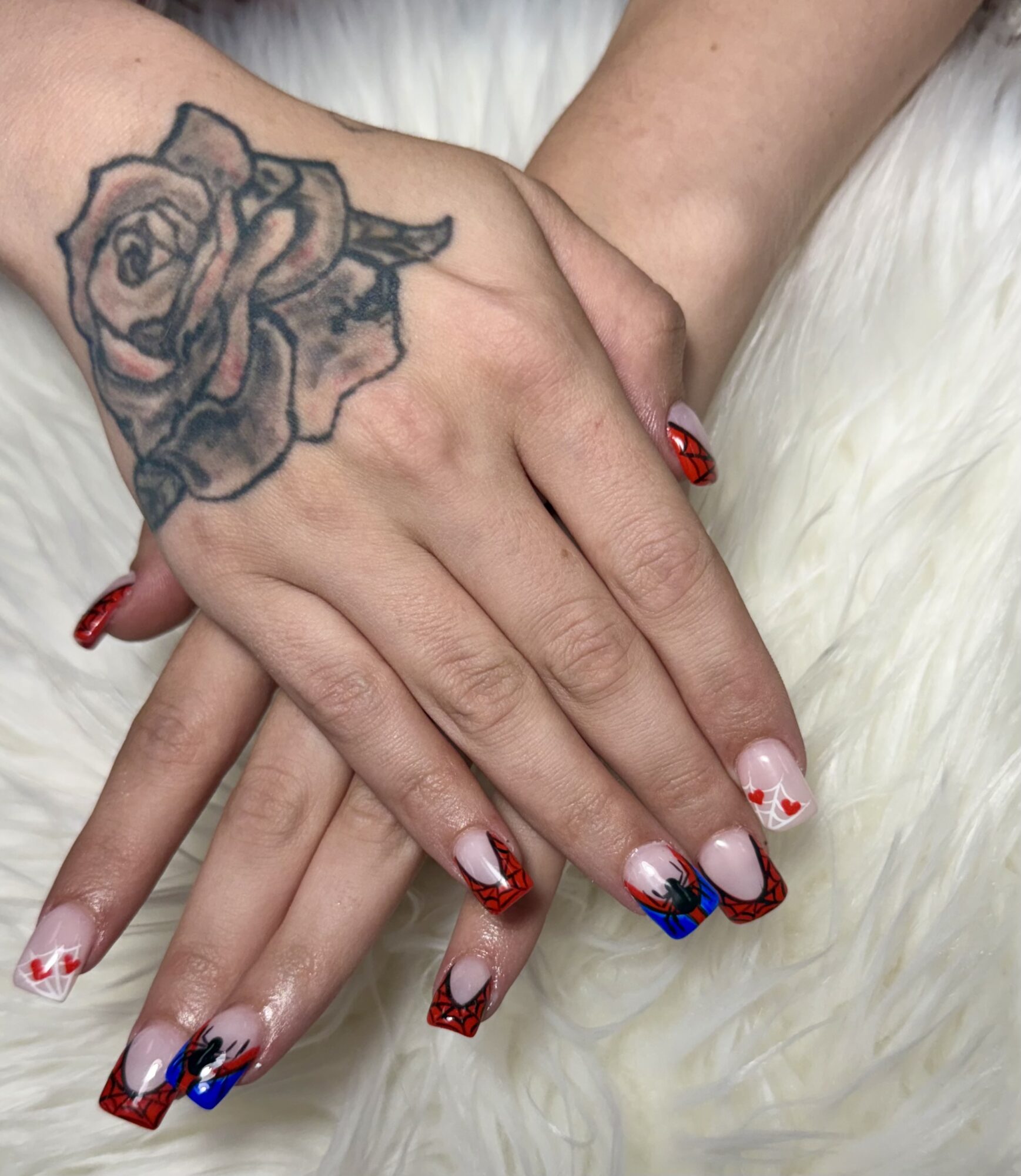 Two hands with decorated nails and a tattoo of a rose on the wrist, resting on a white furry surface.