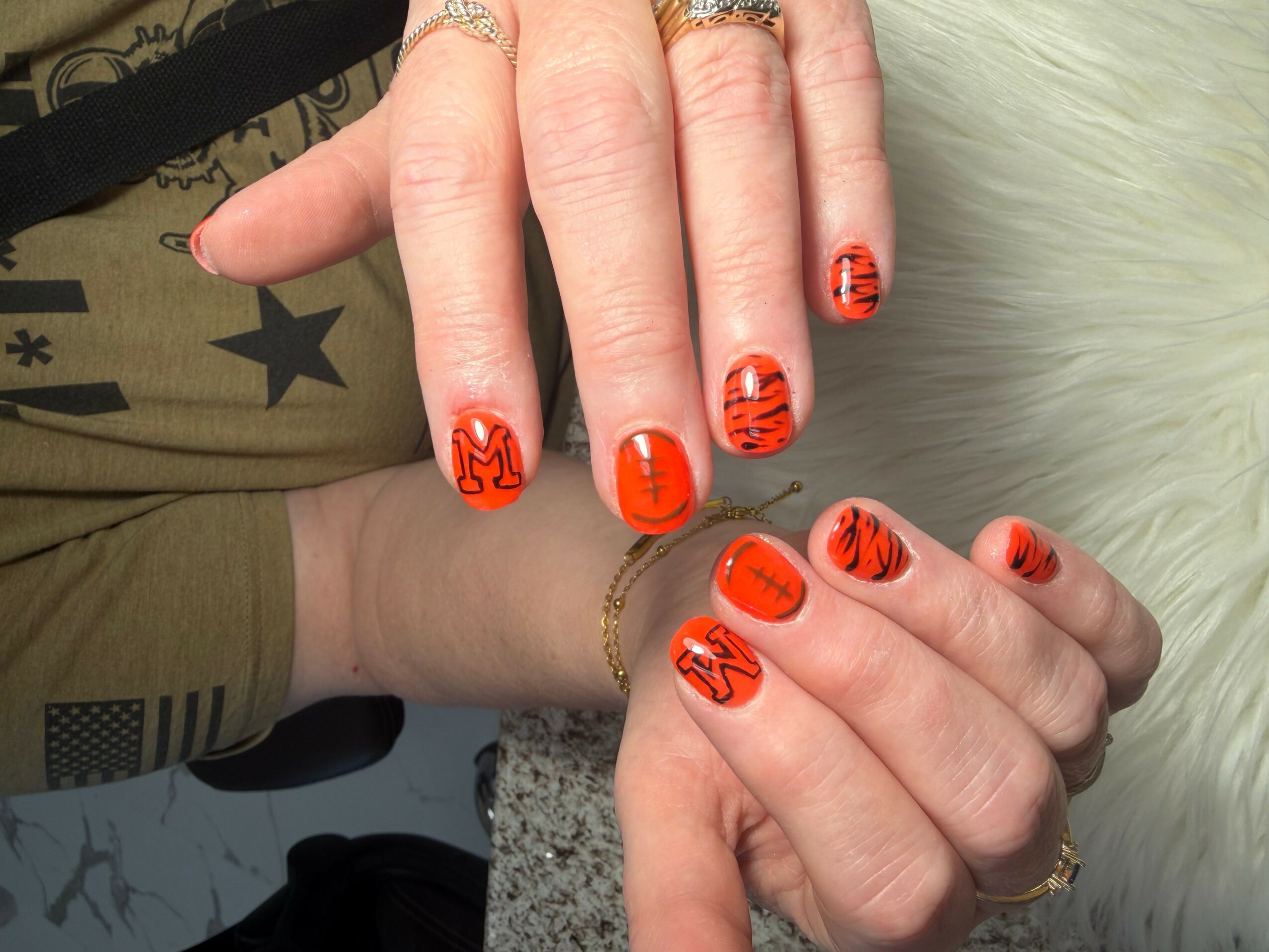 Hands with orange and black striped nail art spelling 'MUSE' and 'LOVE' on fingers, rings, and a bracelet, on a furry surface.