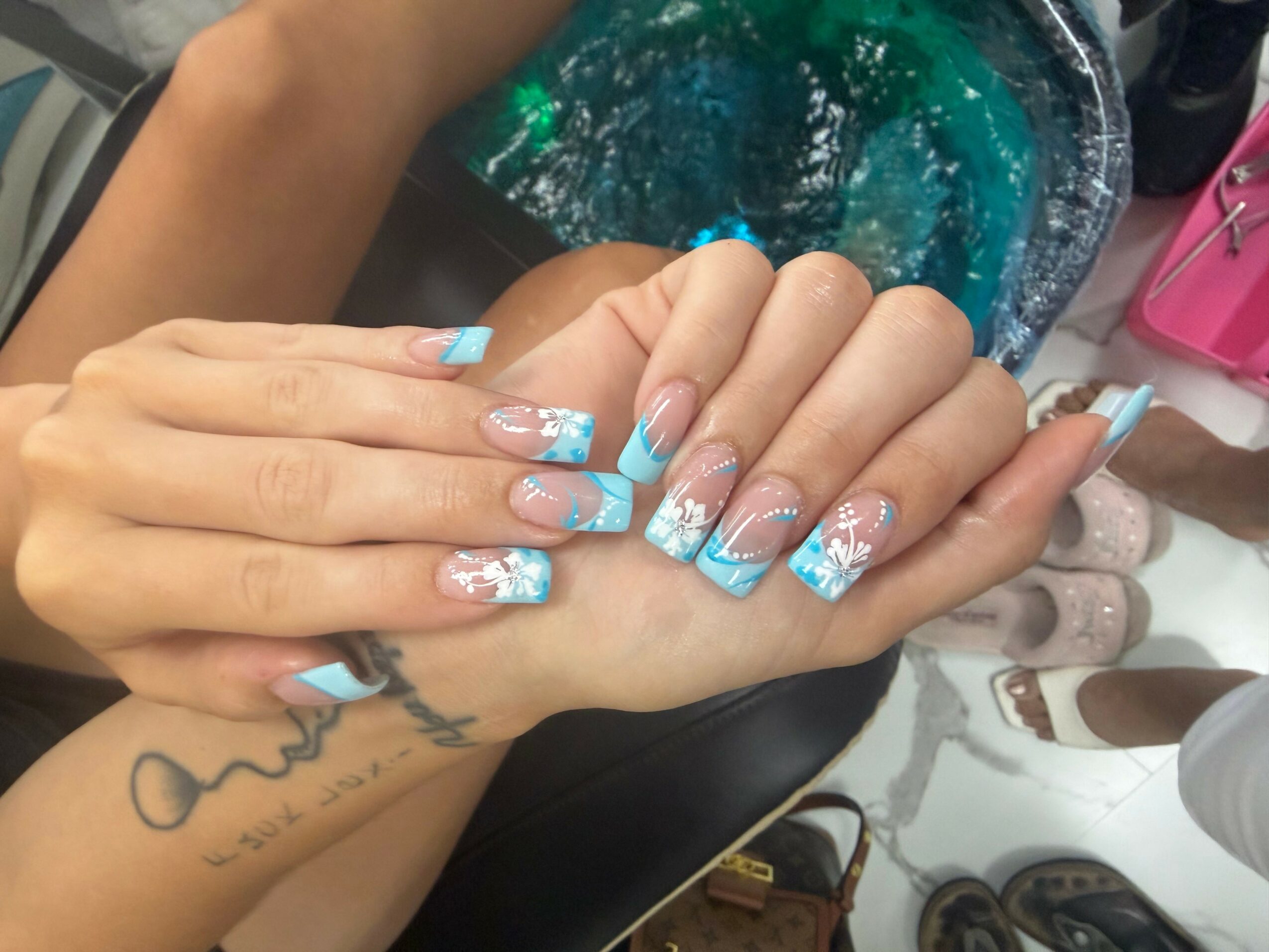 Two hands with decorated nails, one with a tattoo, resting on a surface with a bag and shoes nearby.
