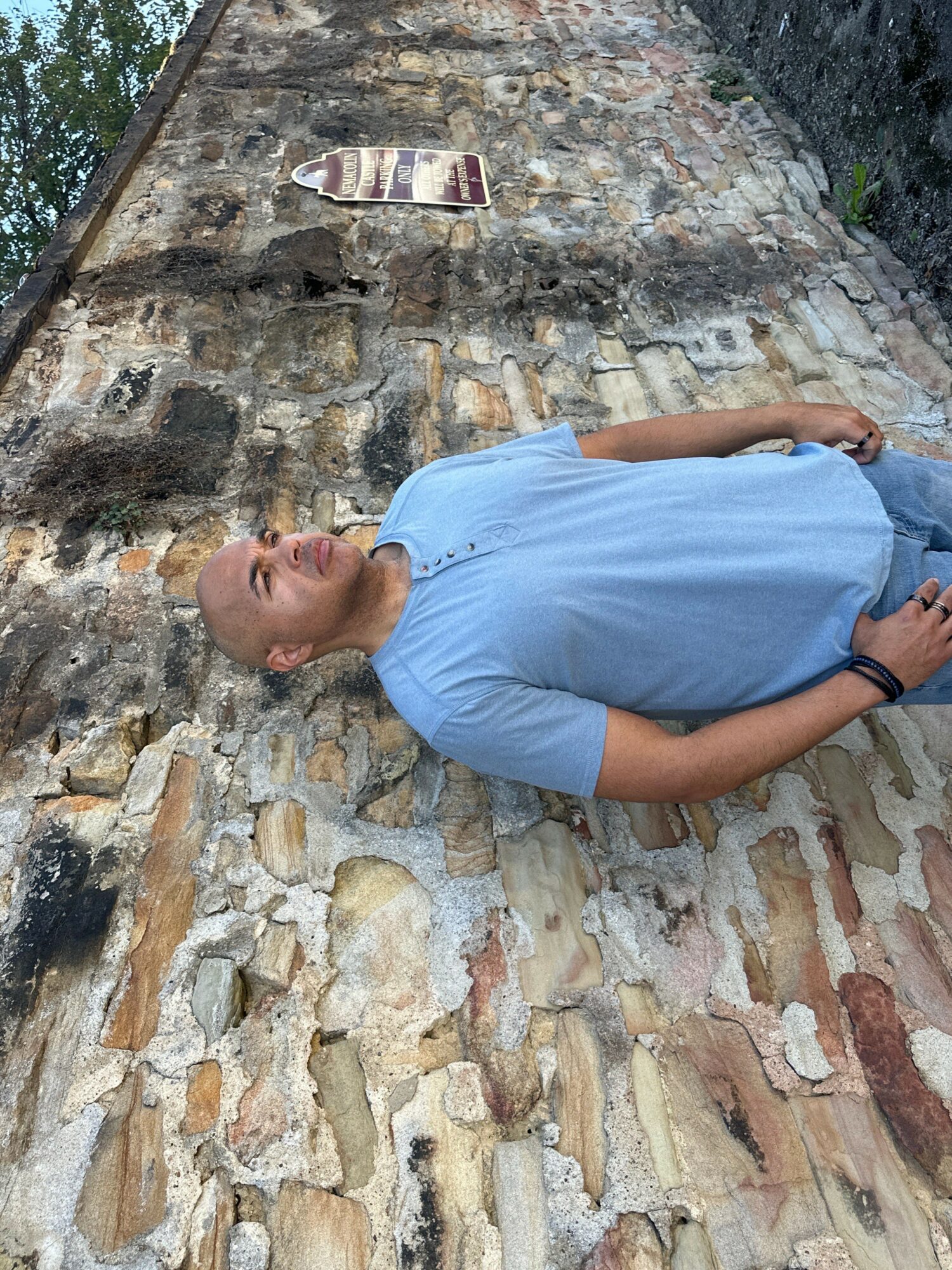 Person standing against a stone wall, wearing a light blue shirt, with arms crossed and head turned to the side.