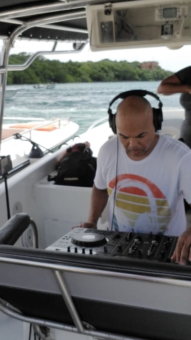 Man wearing headphones and a white t-shirt with a colorful design, operating DJ equipment on boat, water and trees in background.