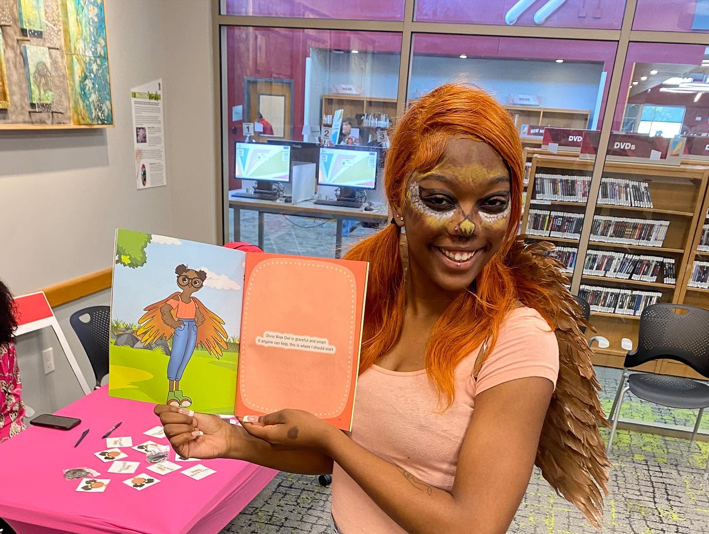 Woman with orange hair holding a children's book inside a library, smiling at the camera.