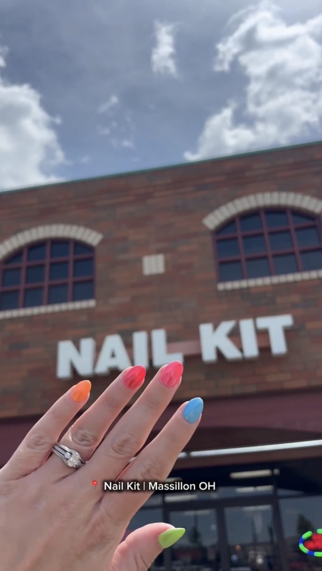 Hand with colorful nails and a ring in front of a brick building with a sign reading 'NAIL KIT'.