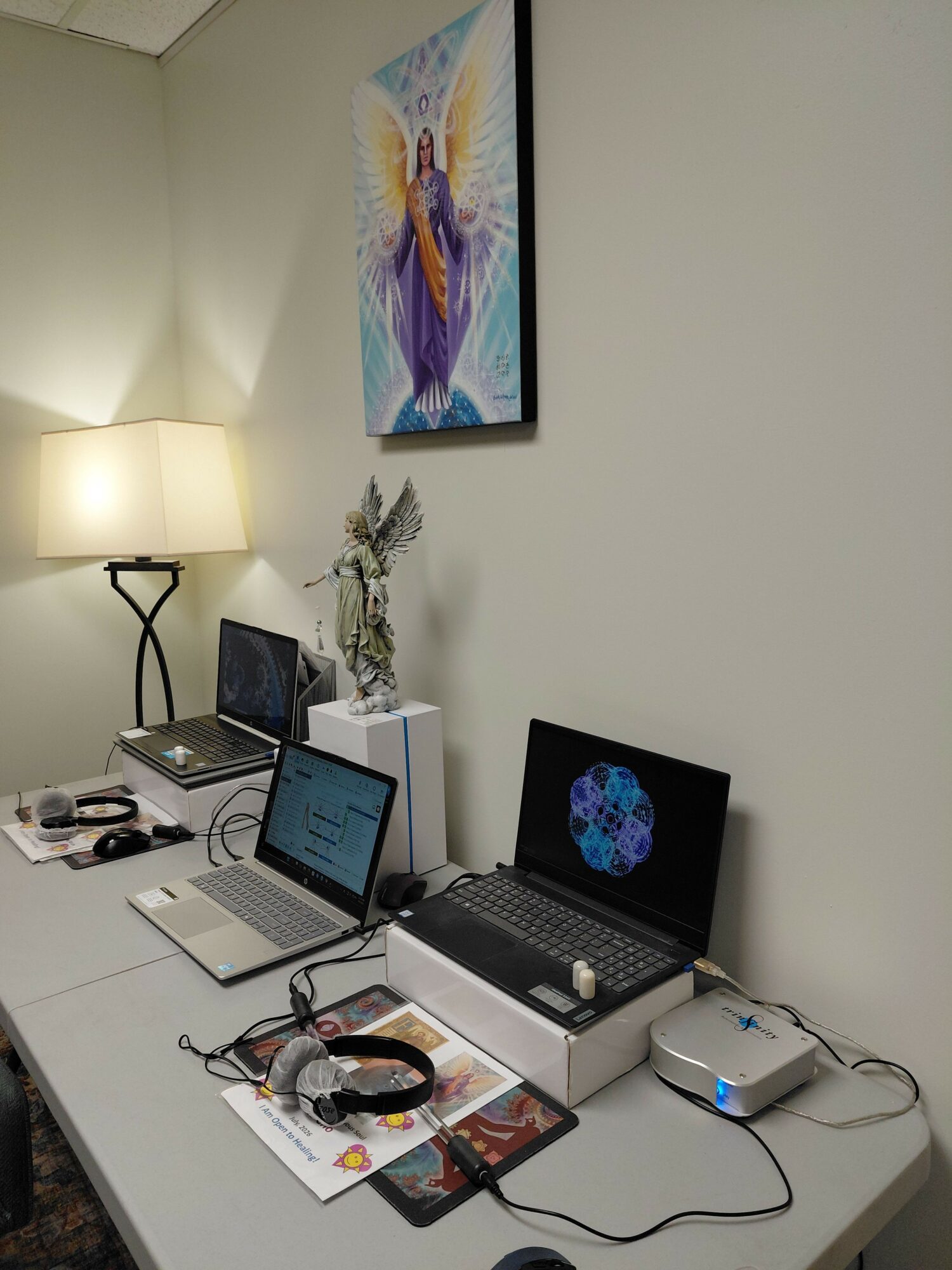 Desk with laptop, tablet, headphones, and framed picture, against a wall with a colorful artwork, illuminated by a lamp.