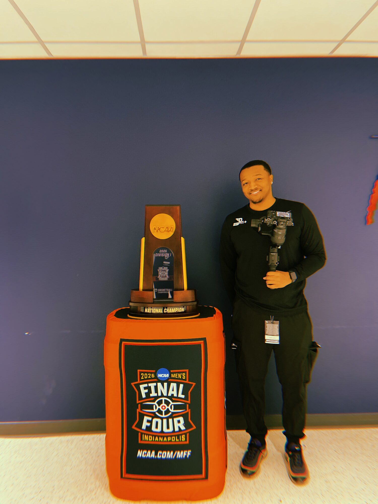Person in black uniform standing next to a trophy display with a Final Four sign, against a blue wall.