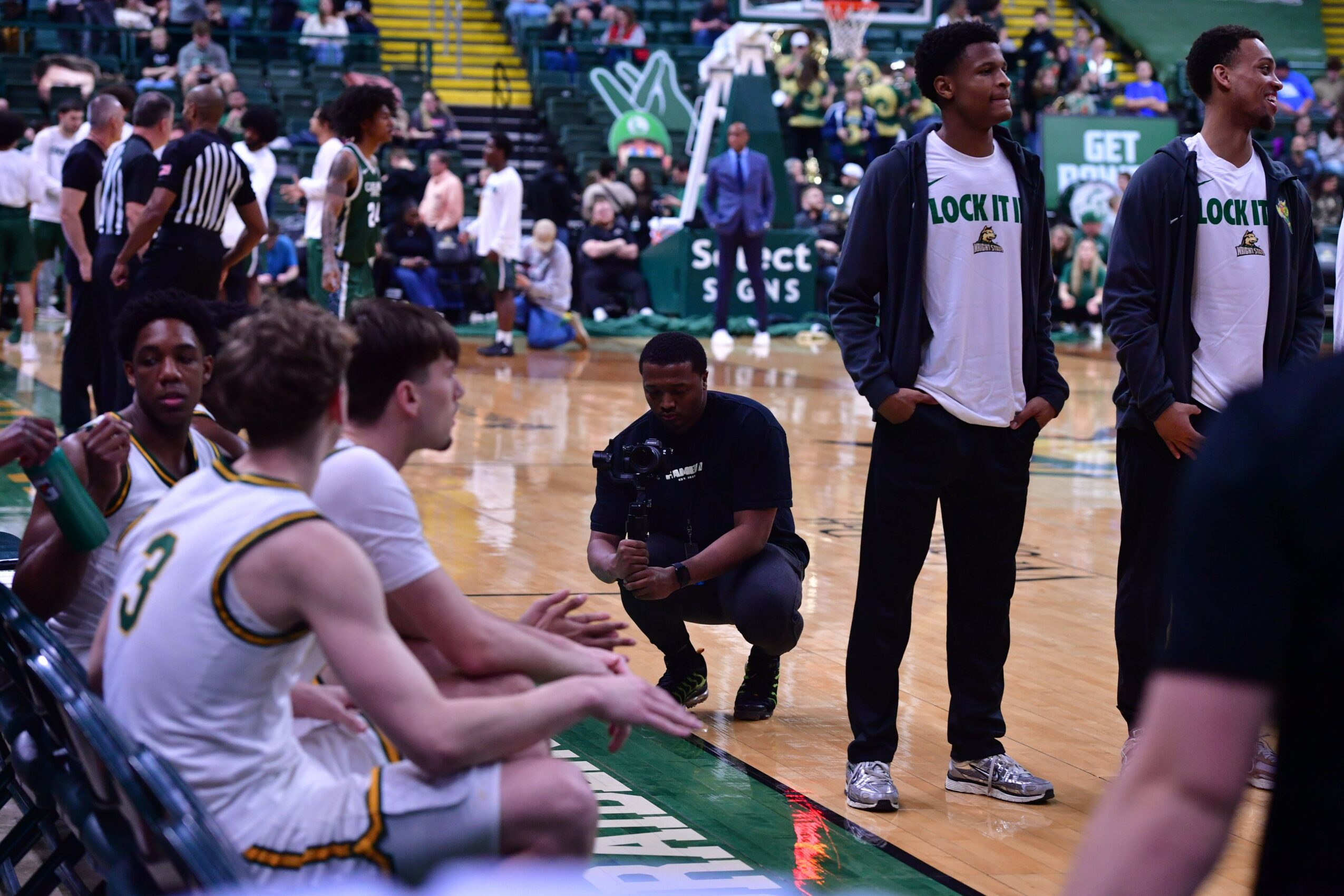 Basketball team seated and standing on court during game, players in white and green uniforms, some sitting, some standing, with spectators in background.